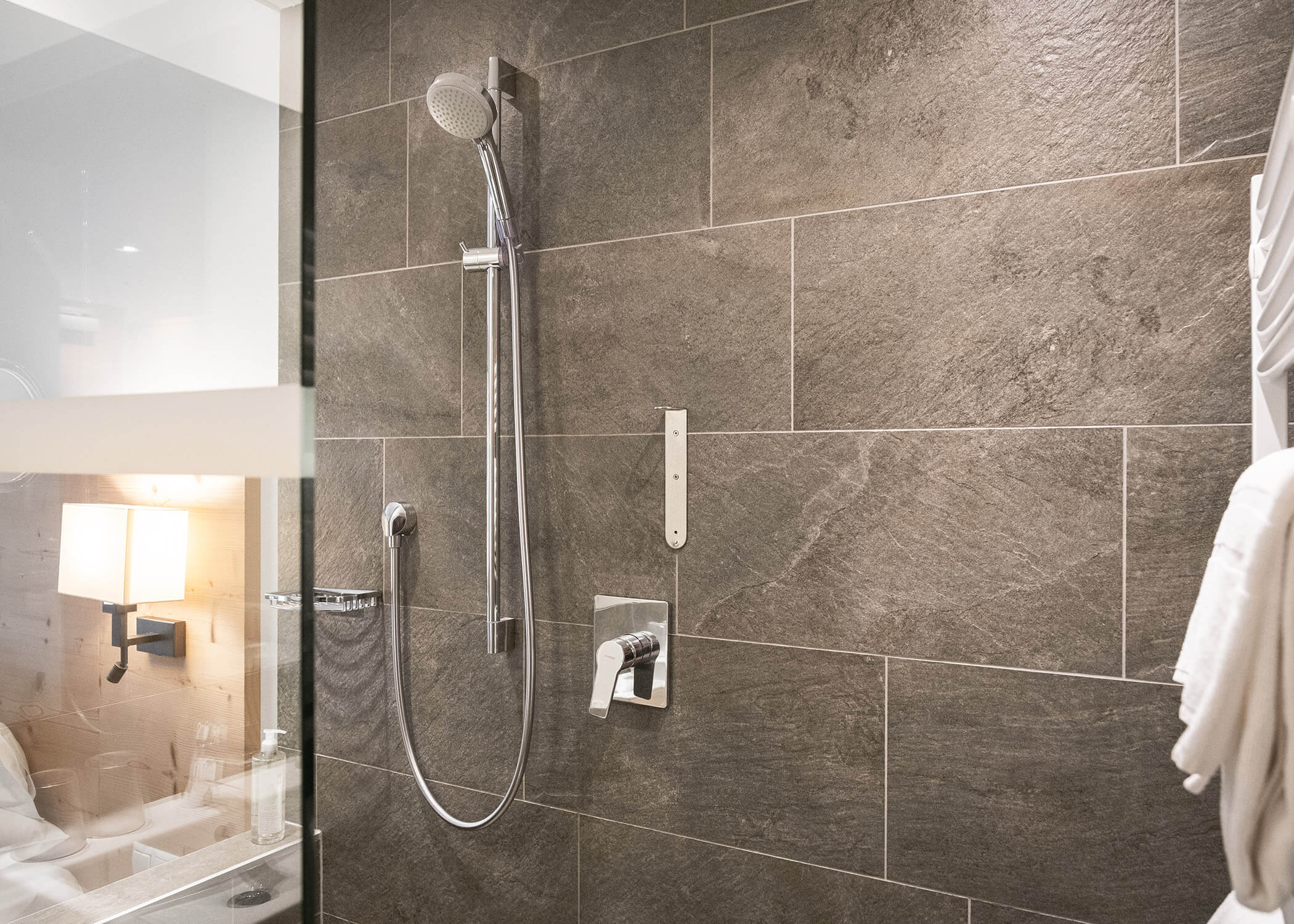 Moderne Dusche mit grau gefliester Wand, Handbrause, Handtuch an einem Haken und Teilansicht eines Badezimmers. - Hotel La Casies Mountain Living