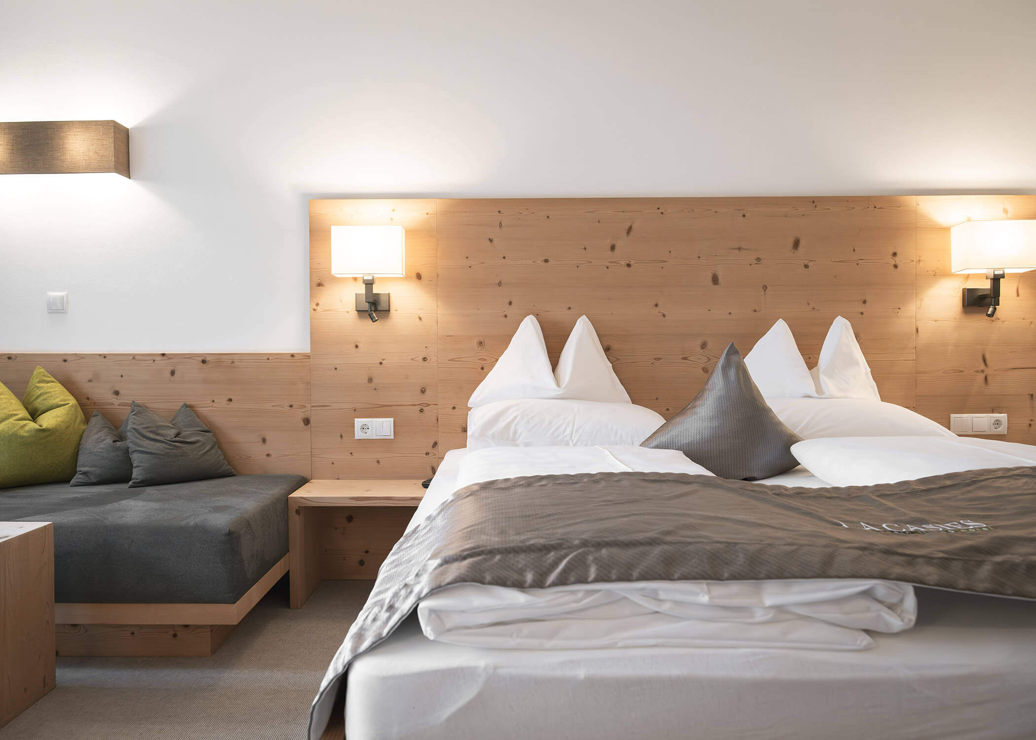 Modernes Hotelzimmer mit Holzkopfteil, Doppelbett, grauer Decke und gemütlicher Sitzecke mit grünen Kissen. - Hotel La Casies Mountain Living