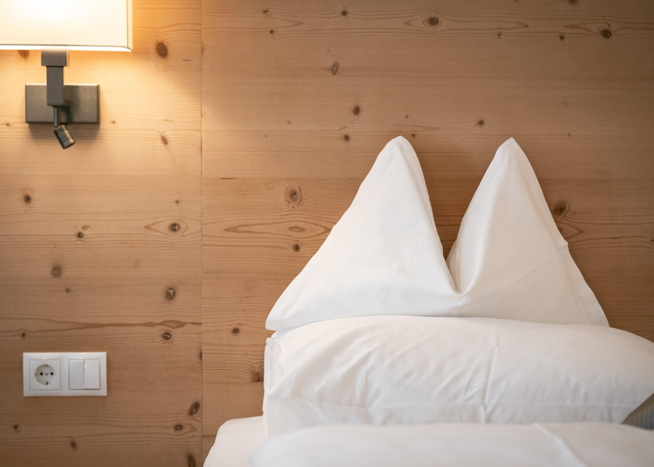 Zwei weiße, zu Dreiecken gefaltete Kissen auf einem Bett mit einer Holzwand, einer Lampe und einer Steckdose in der Nähe. - Hotel La Casies Mountain Living