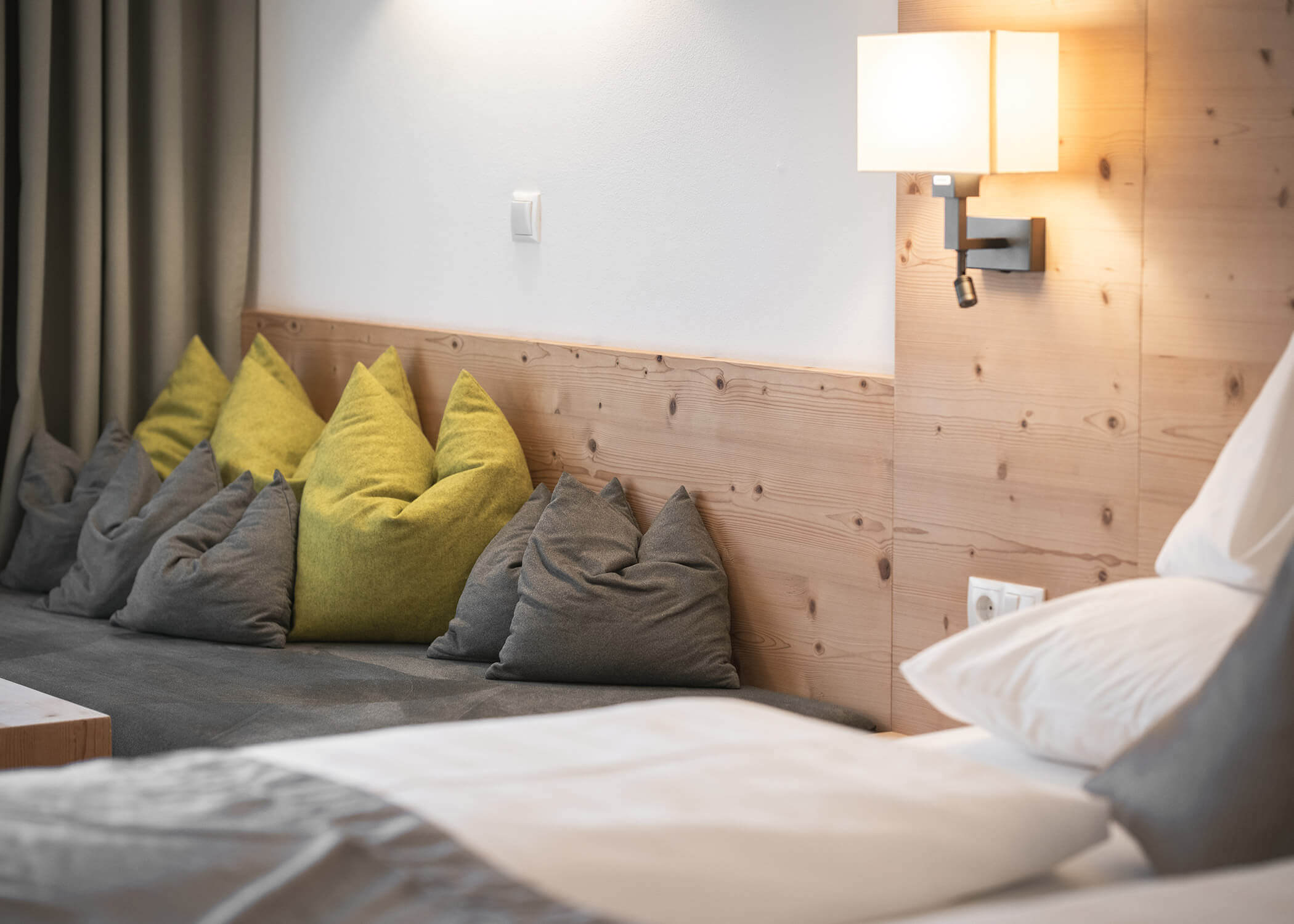 Gemütliches Schlafzimmer mit einem Bett, grauen und grünen Kissen, einem Kopfteil aus Holz und einer Wandlampe. - Hotel La Casies Mountain Living