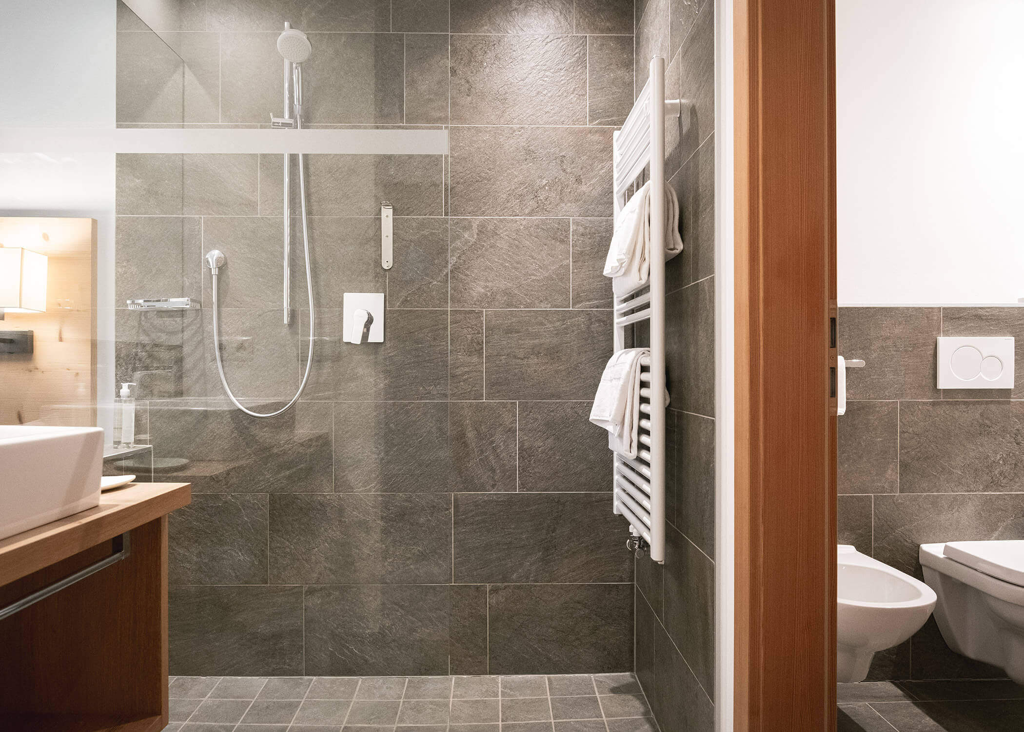 Modernes Badezimmer mit grau gefliesten Wänden, begehbarer Dusche, Handtuchhalter, Waschbecken und Toilette. - Hotel La Casies Mountain Living