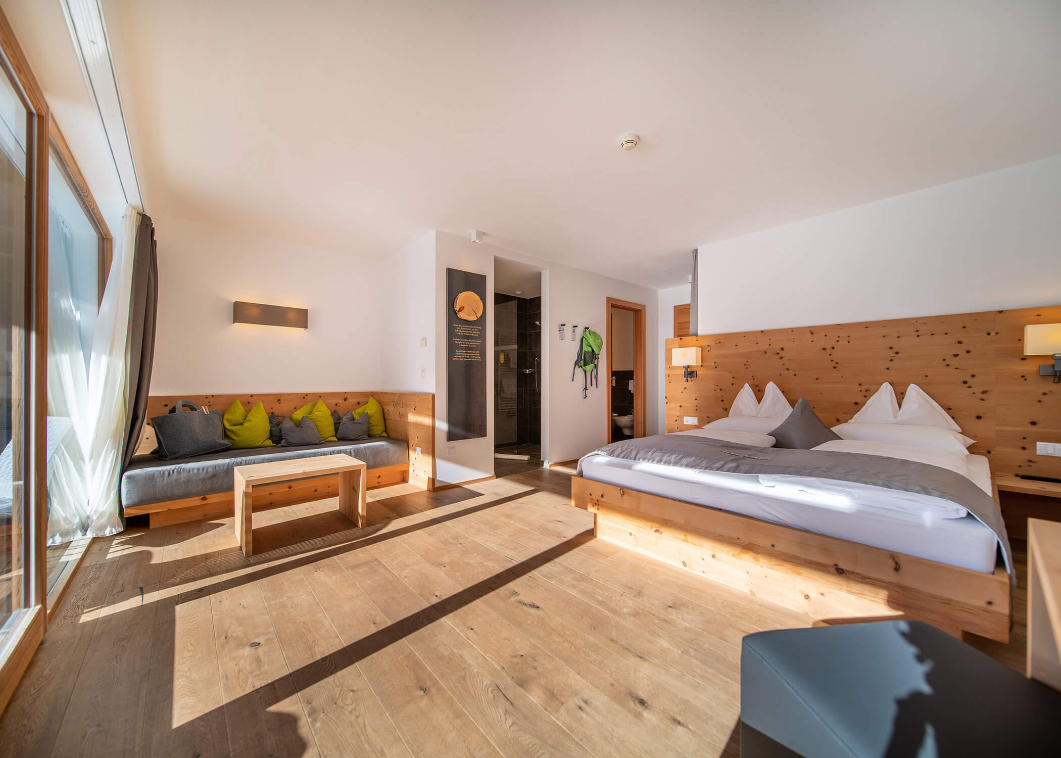 Helles Hotelzimmer mit Holzböden, einem großen Bett, einem Sofa und Sonnenlicht, das durch große Fenster einfällt. - Hotel La Casies Mountain Living