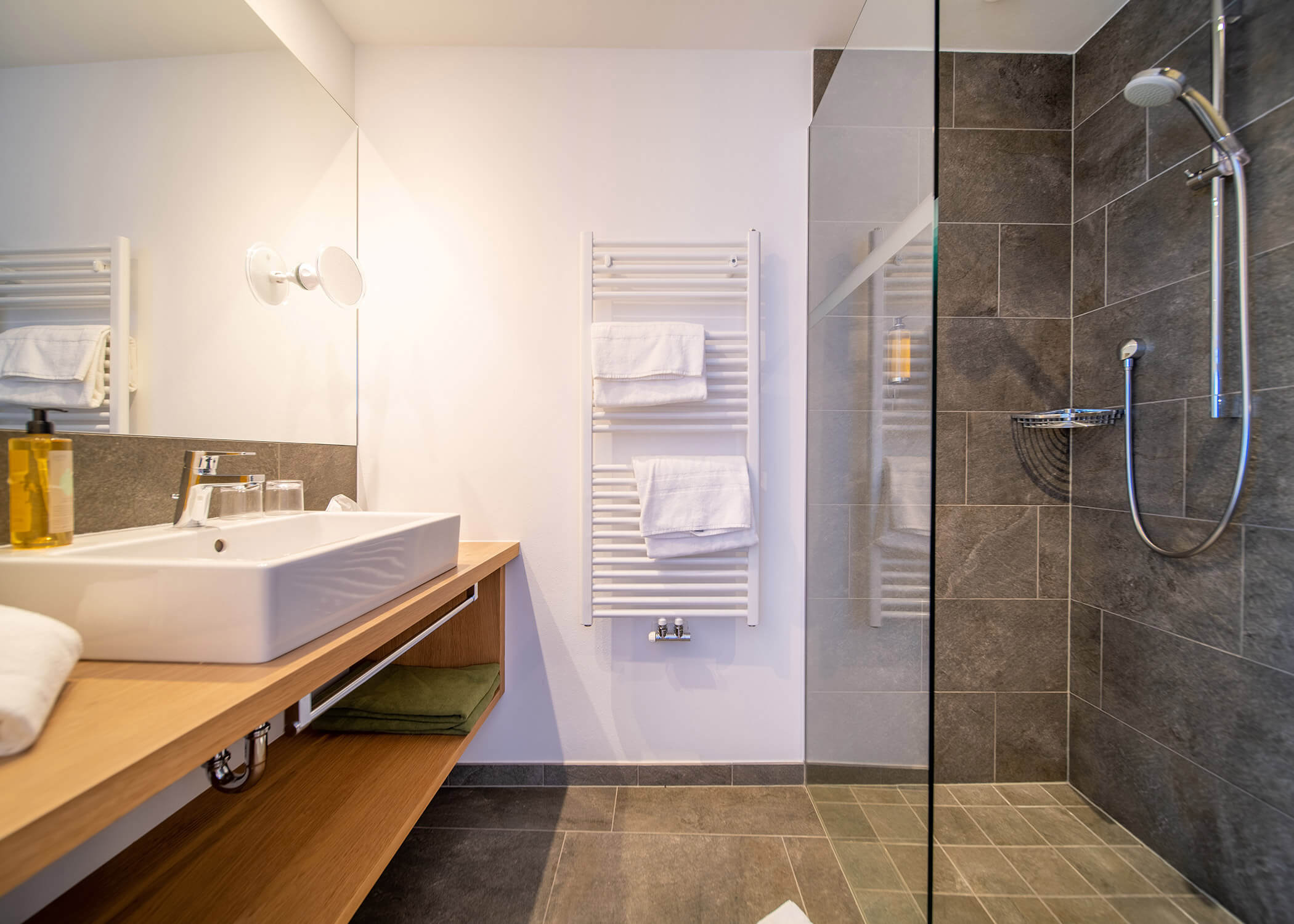 Modernes Badezimmer mit Waschbecken, Holzarbeitsplatte, Handtuchhalter und begehbarer Dusche mit grauen Fliesen. - Hotel La Casies Mountain Living
