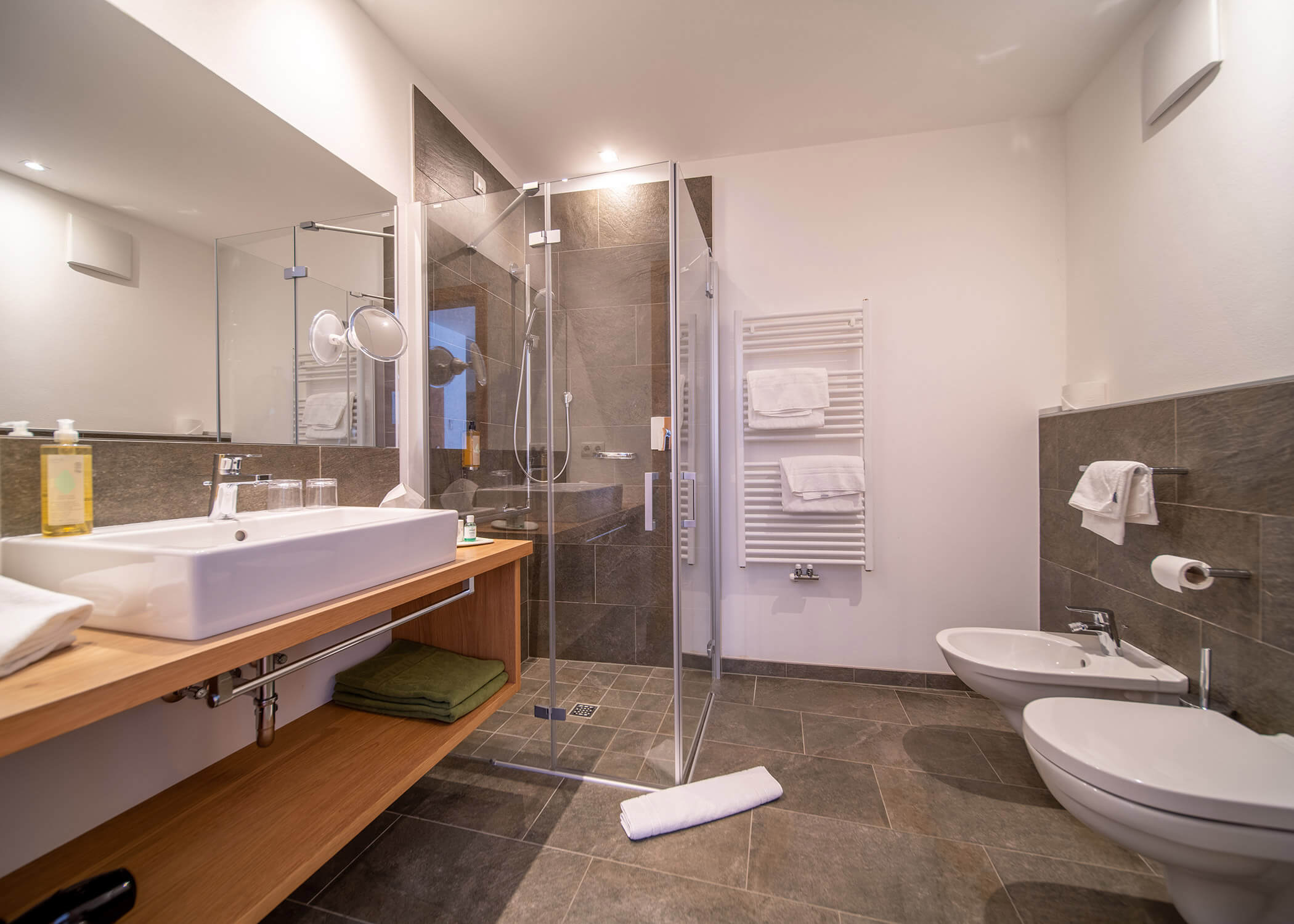 Modernes Badezimmer mit Glasdusche, Waschbecken, Handtuchhalter, Bidet, Toilette und grau gefliestem Boden und Wänden. - Hotel La Casies Mountain Living