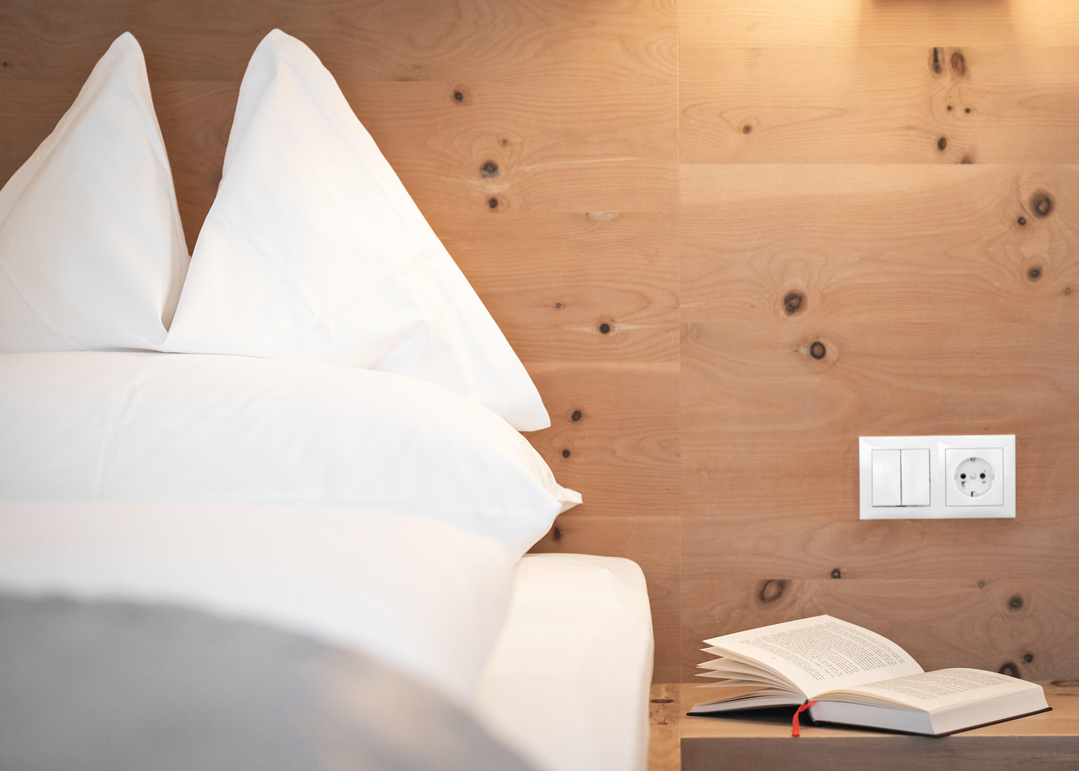 Ein ordentlich gemachtes Bett mit weißen Kissen neben einem aufgeschlagenen Buch auf einem hölzernen Nachttisch neben einer Steckdose. - Hotel La Casies Mountain Living