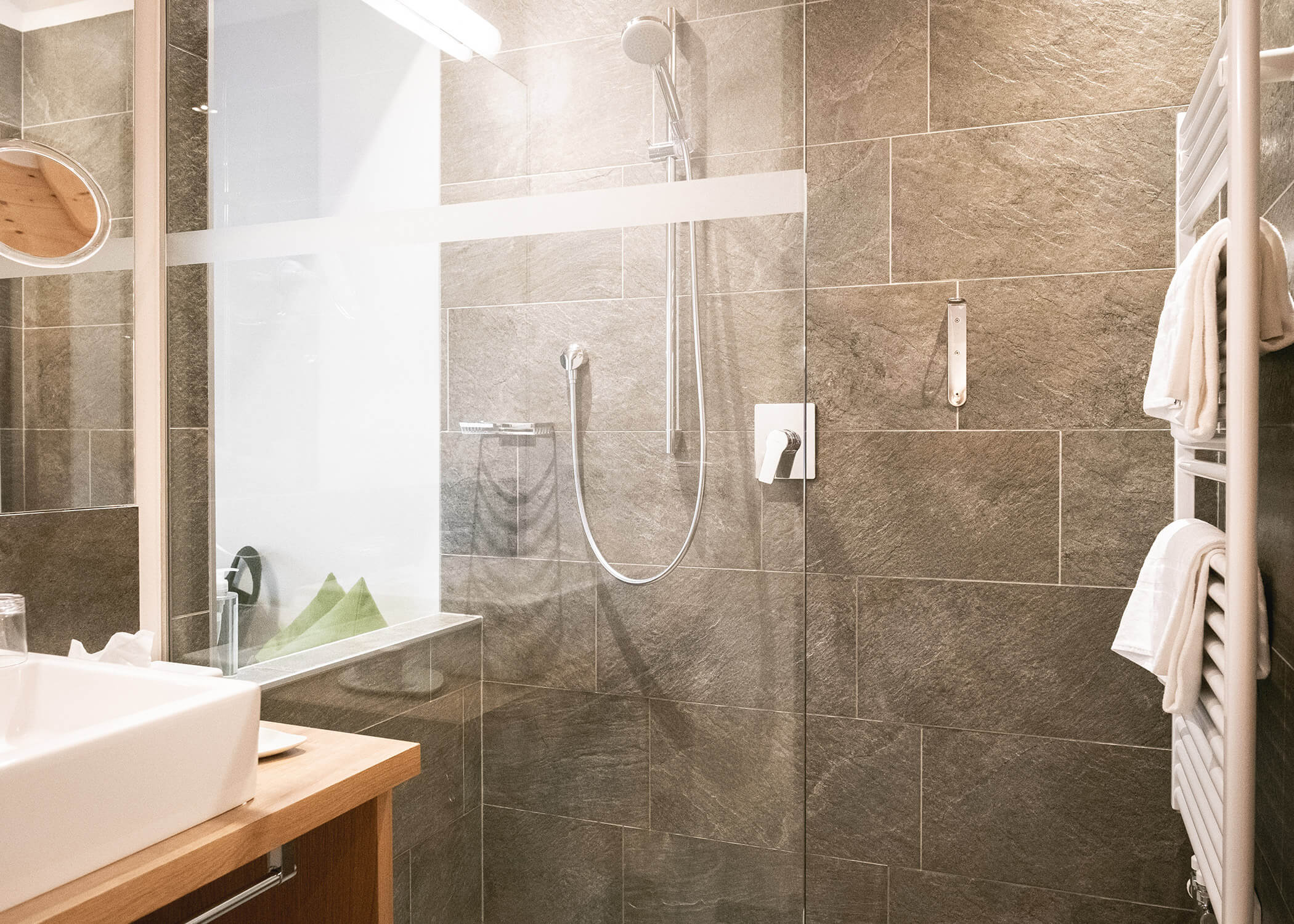 Modernes Badezimmer mit einer Glasdusche, Steinfliesenwänden, weißem Waschbecken und Handtüchern, die auf einem Ständer hängen. - Hotel La Casies Mountain Living