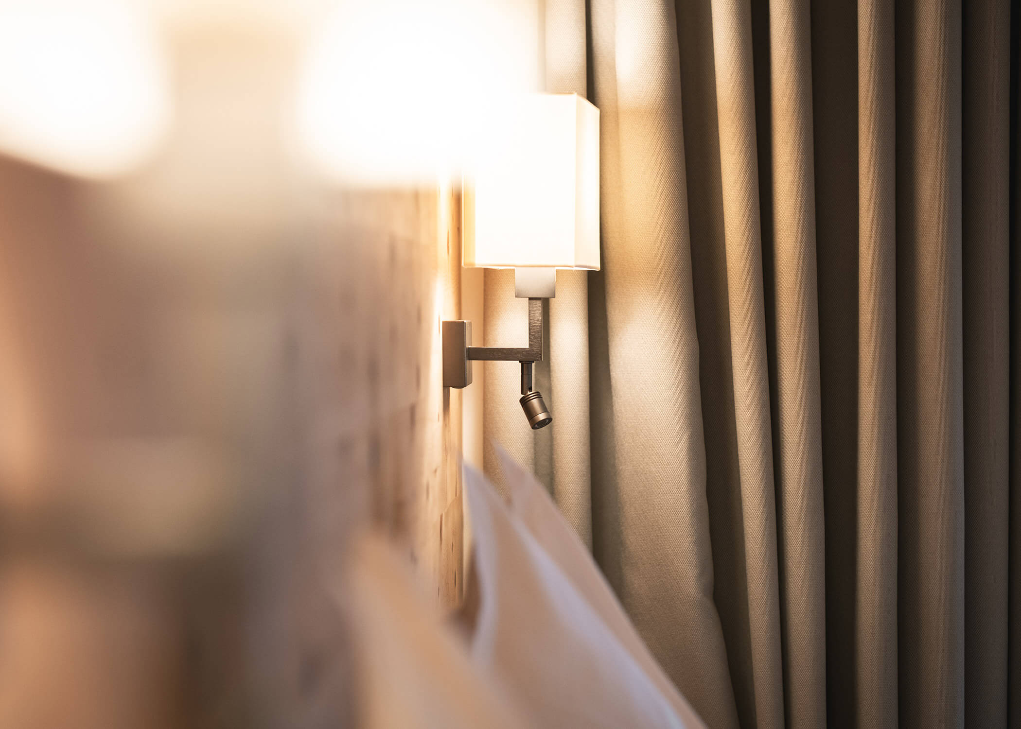 Wandlampe neben einem Bett mit beigen Vorhängen im Hintergrund, die eine gemütliche Atmosphäre schaffen. - Hotel La Casies Mountain Living