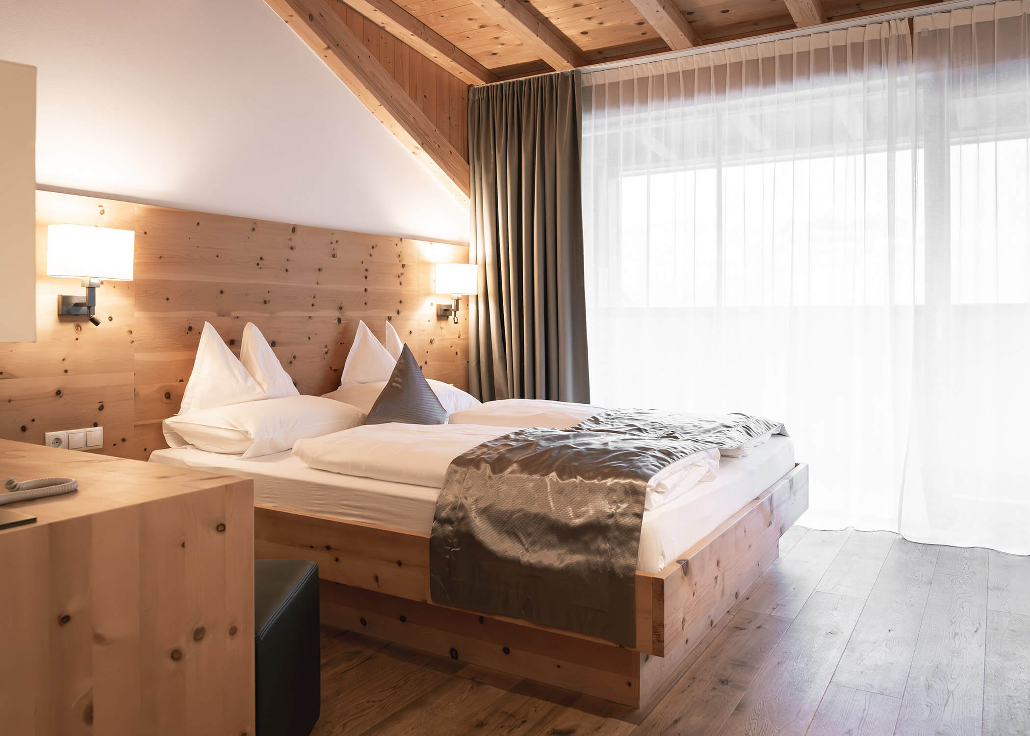 Gemütliches Hotelzimmer mit Holzwänden, Doppelbett, Nachttischlampen, Schreibtisch und großem Fenster mit durchsichtigen Vorhängen. - Hotel La Casies Mountain Living