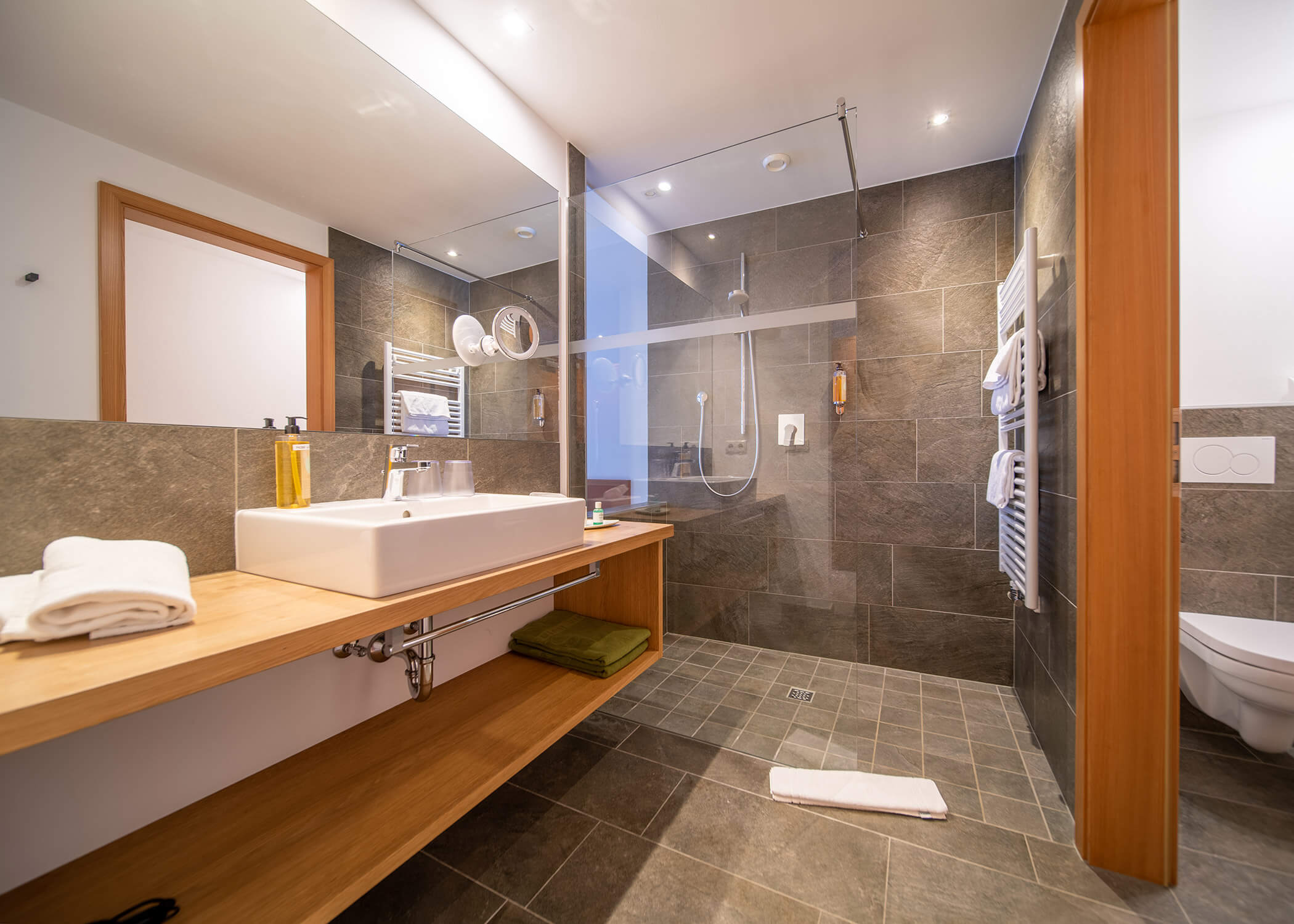 Modernes Badezimmer mit begehbarer Dusche, großem Spiegel, Waschbecken, Handtüchern und Holzregalen. - Hotel La Casies Mountain Living