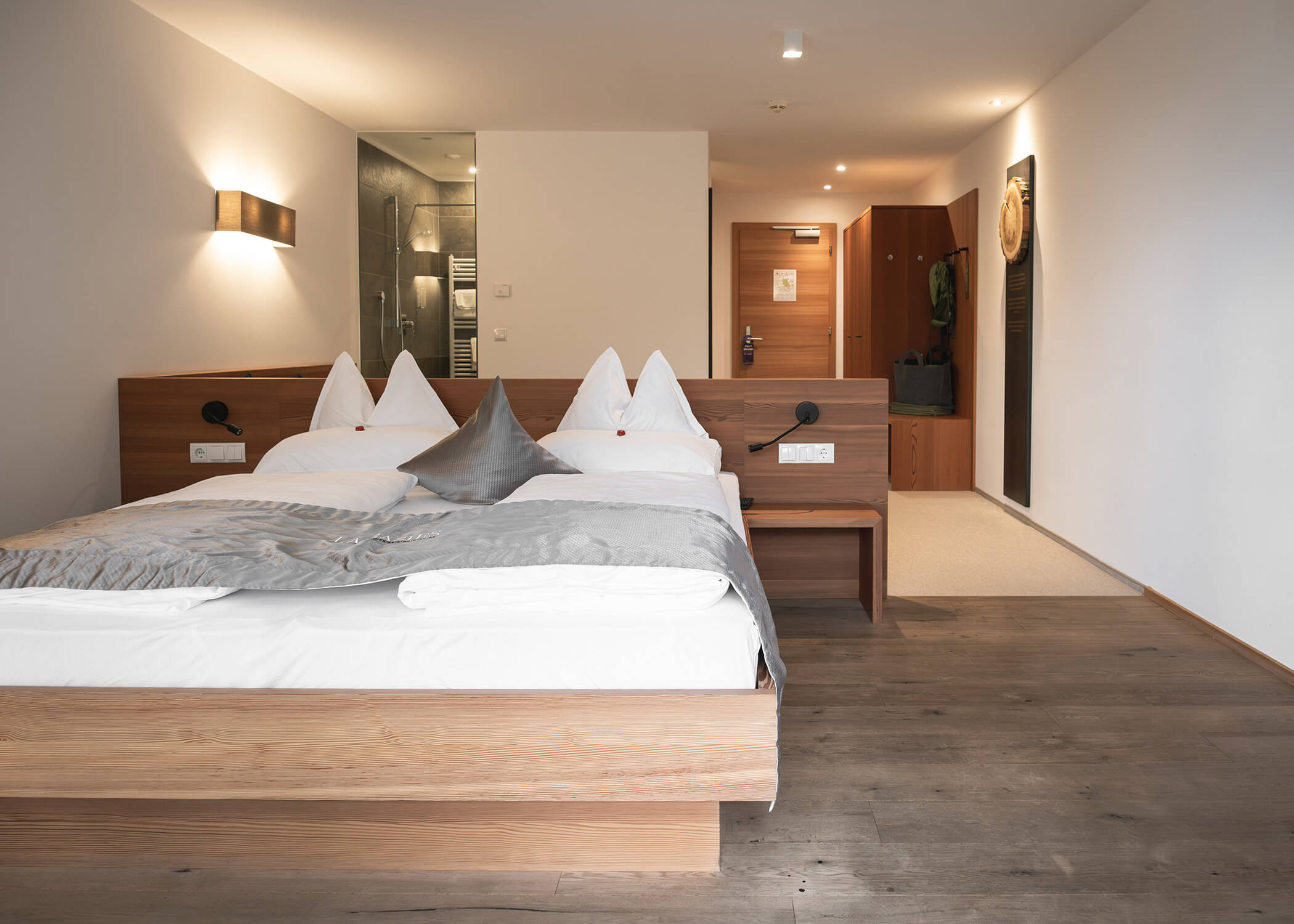 Modernes Hotelzimmer mit Holzbett, grauer Bettwäsche und Blick auf den Eingangs- und Badbereich. - Hotel La Casies Mountain Living