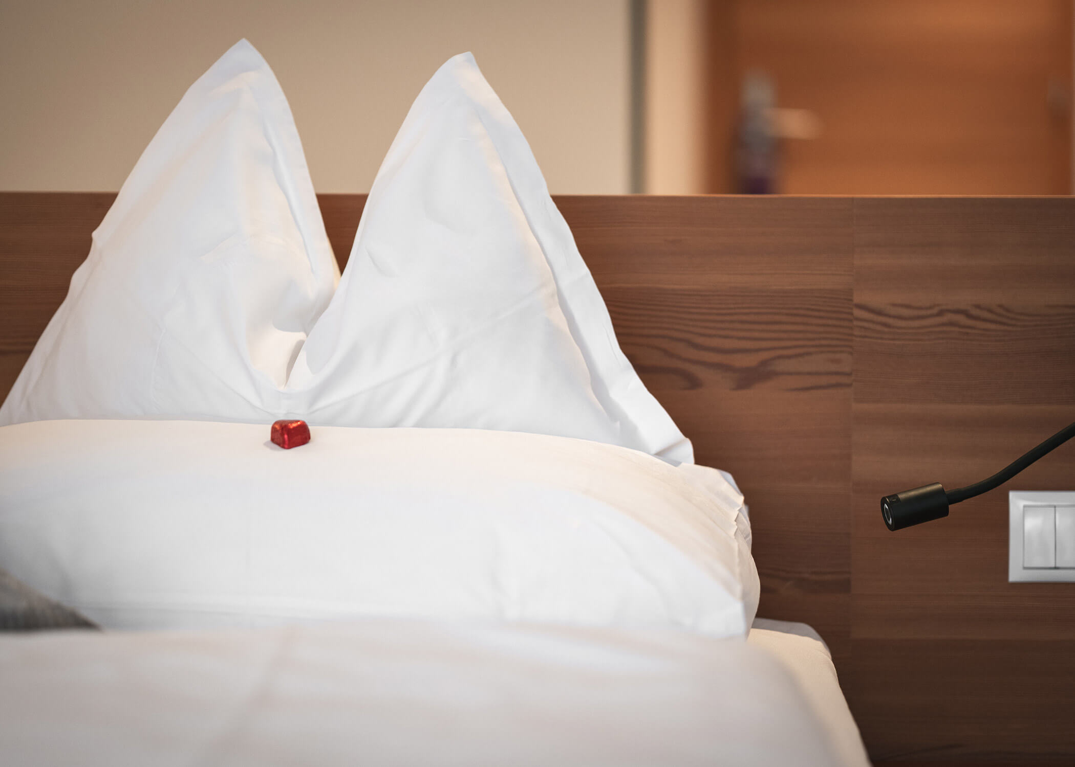 Ein ordentlich gemachtes Bett mit weißen Kissen und einer roten Schokolade darauf, daneben ein hölzernes Kopfteil. - Hotel La Casies Mountain Living