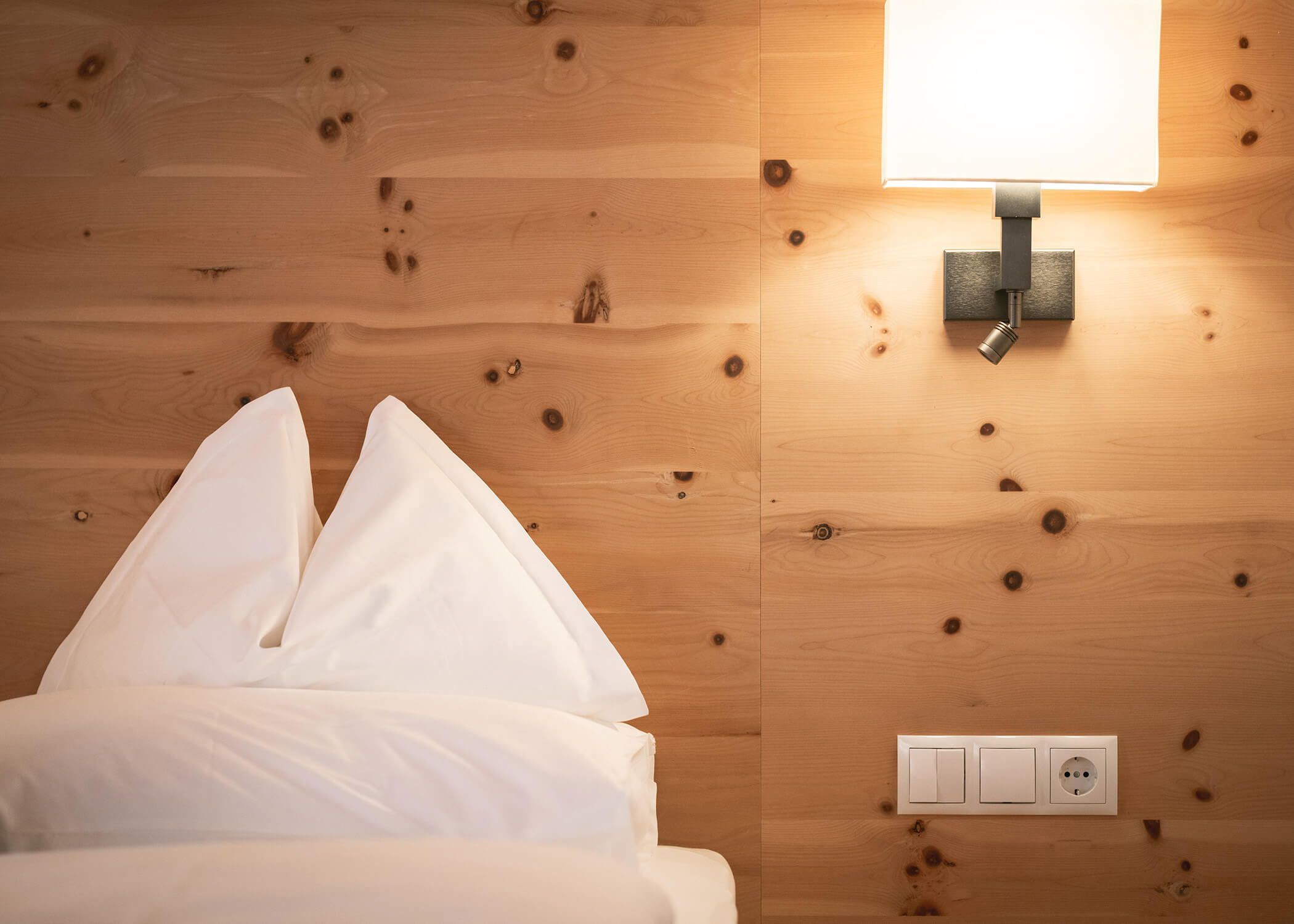 Weiße Kissen auf einem ordentlich gemachten Bett neben einer Holzwand mit einer Lampe und Steckdosen. - Hotel La Casies Mountain Living