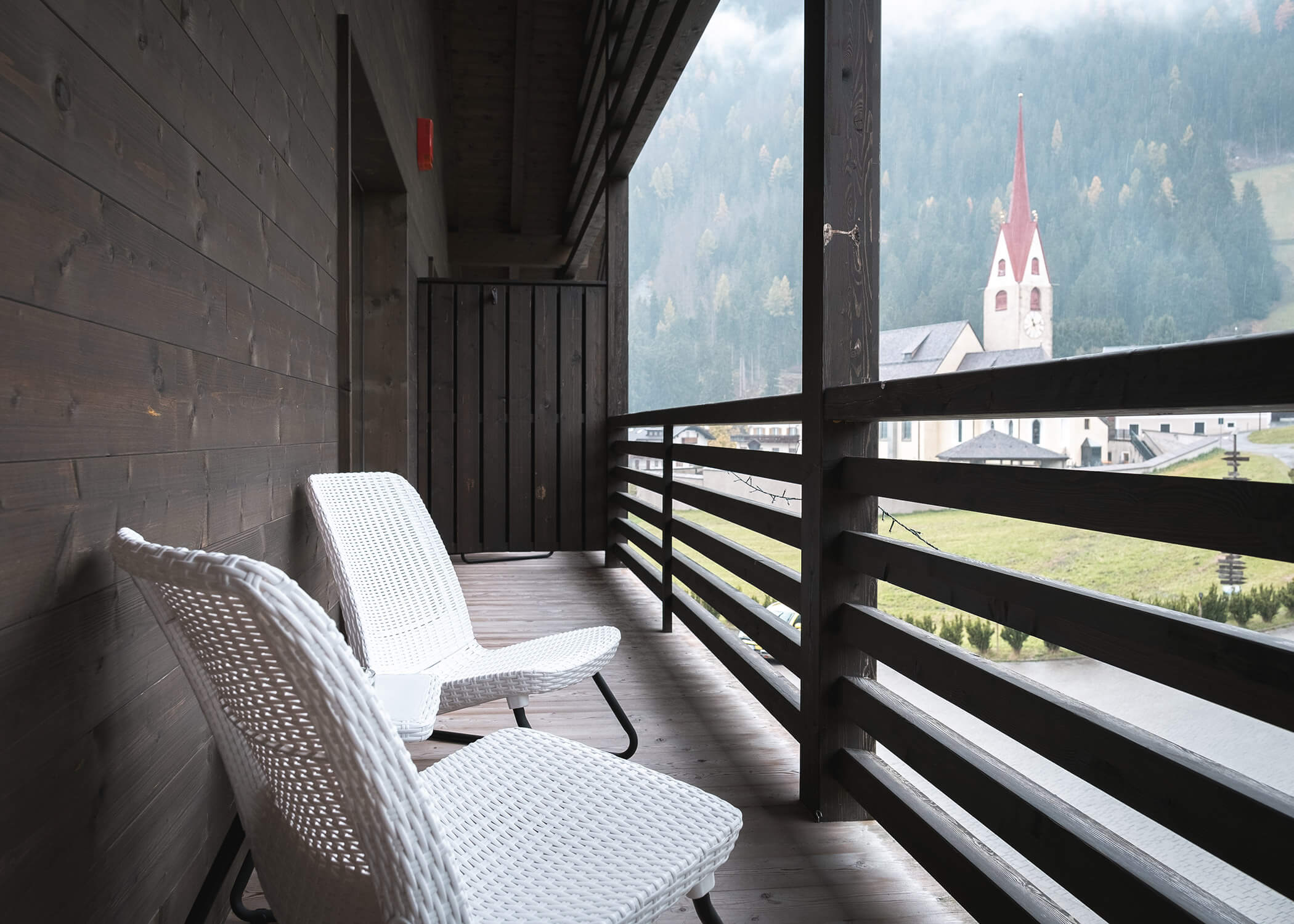 Zwei weiße Stühle auf einem Holzbalkon mit Blick auf ein Dorf mit einer Kirche und Bergen im Hintergrund. - Hotel La Casies Mountain Living