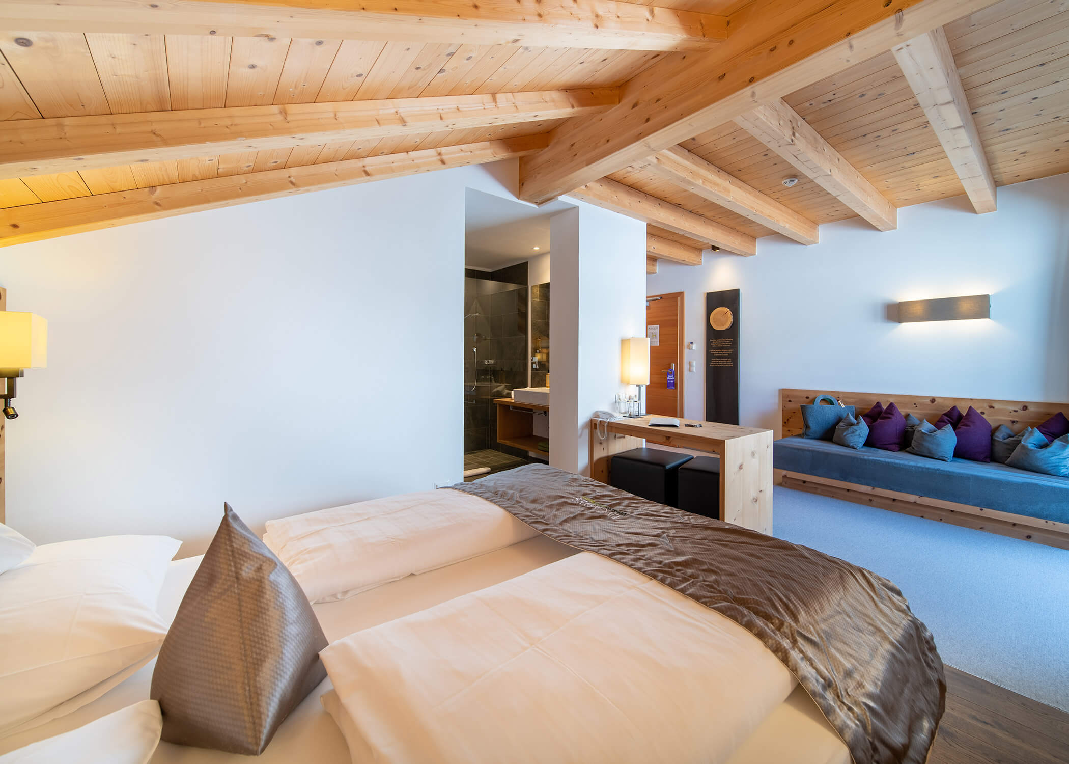 Modernes Hotelzimmer mit Holzdecke, Doppelbett, Couch mit bunten Kissen und offenem Badbereich. - Hotel La Casies Mountain Living