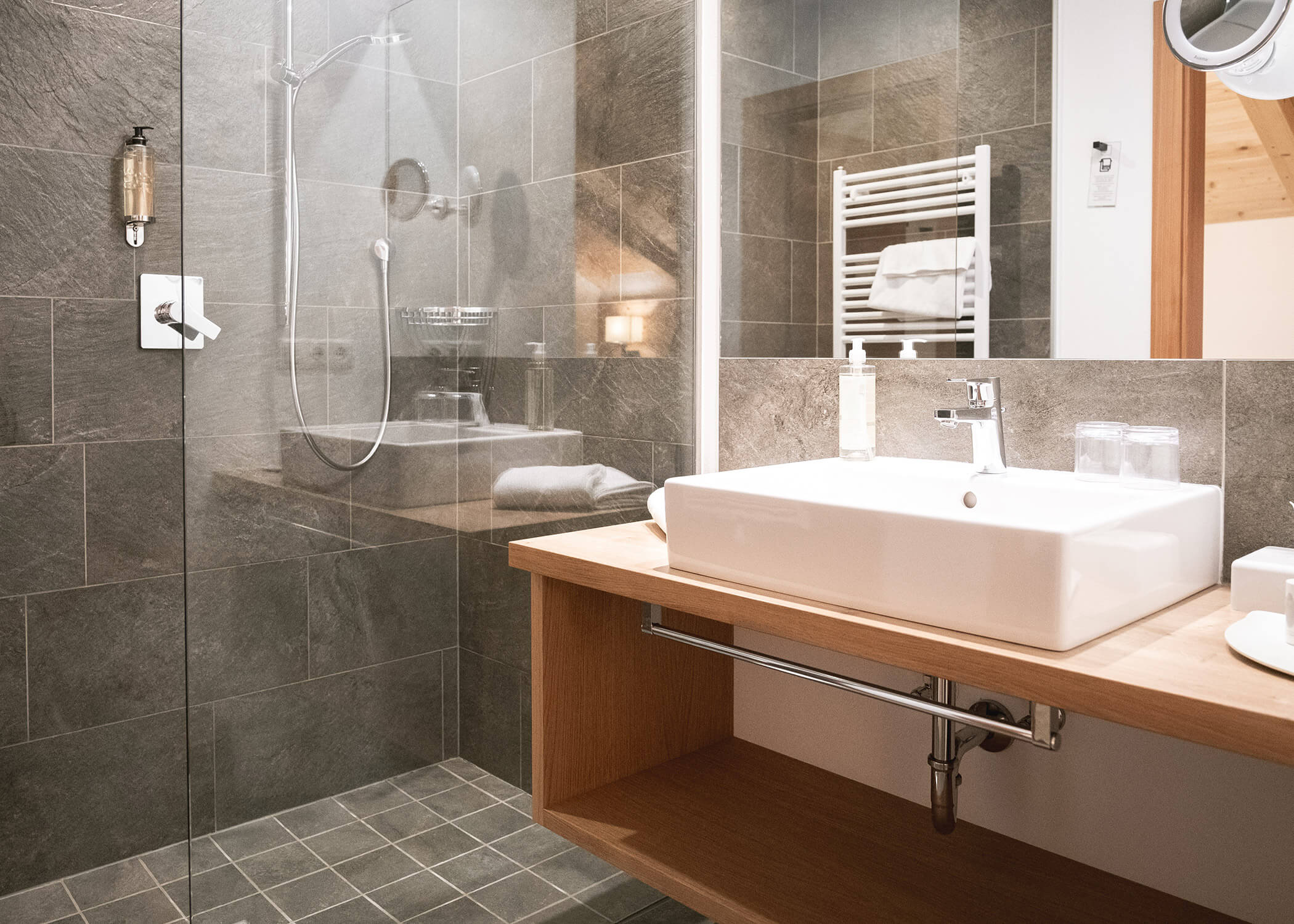 Modernes Badezimmer mit begehbarer Dusche, grauen Fliesen, weißem Waschbecken und einem hölzernen Waschtisch mit Handtüchern. - Hotel La Casies Mountain Living