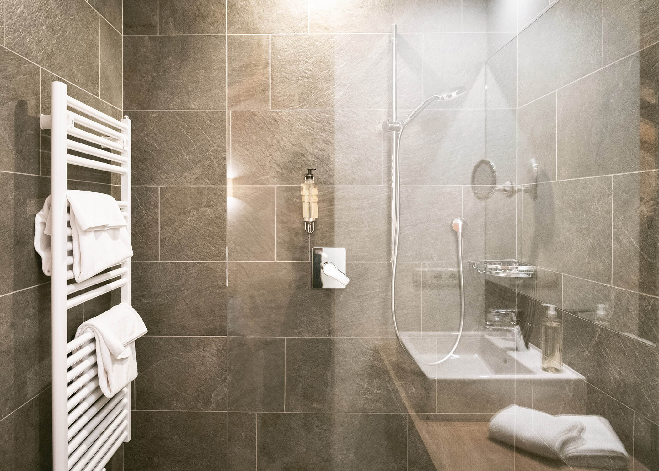 Modernes Badezimmer mit grau gefliesten Wänden, einem Handtuchwärmer, einer Glasdusche und ordentlich gefalteten weißen Handtüchern. - Hotel La Casies Mountain Living