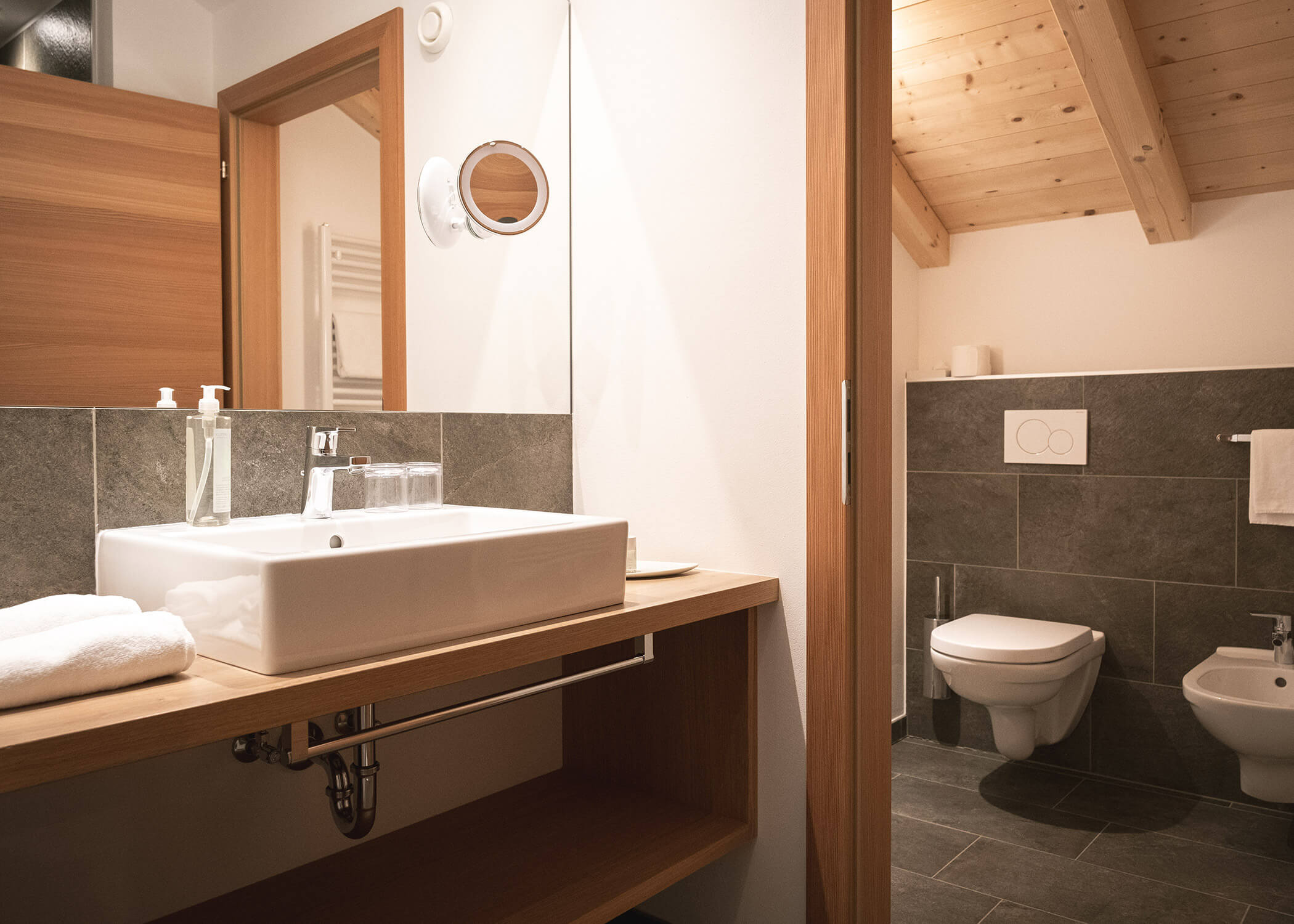 Modernes Badezimmer mit weißem Waschbecken, Spiegel, Toilette, Bidet und Holzakzenten unter einer schrägen Decke. - Hotel La Casies Mountain Living