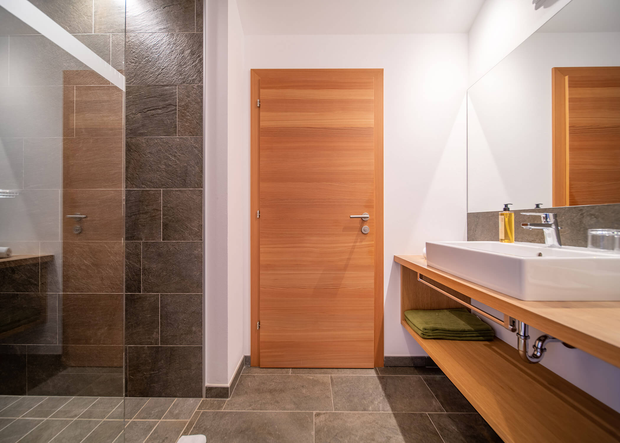 Modernes Badezimmer mit Holztür, großem Spiegel, Waschbecken und einer Dusche mit Glasabtrennung und grauen Fliesen. - Hotel La Casies Mountain Living