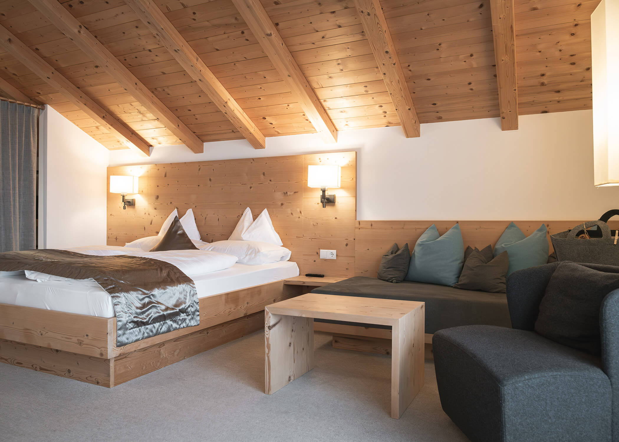 Gemütliches Holzschlafzimmer mit Doppelbett, weichen Kissen, einem kleinen Tisch und einer grauen Couch unter einer schrägen Decke. - Hotel La Casies Mountain Living