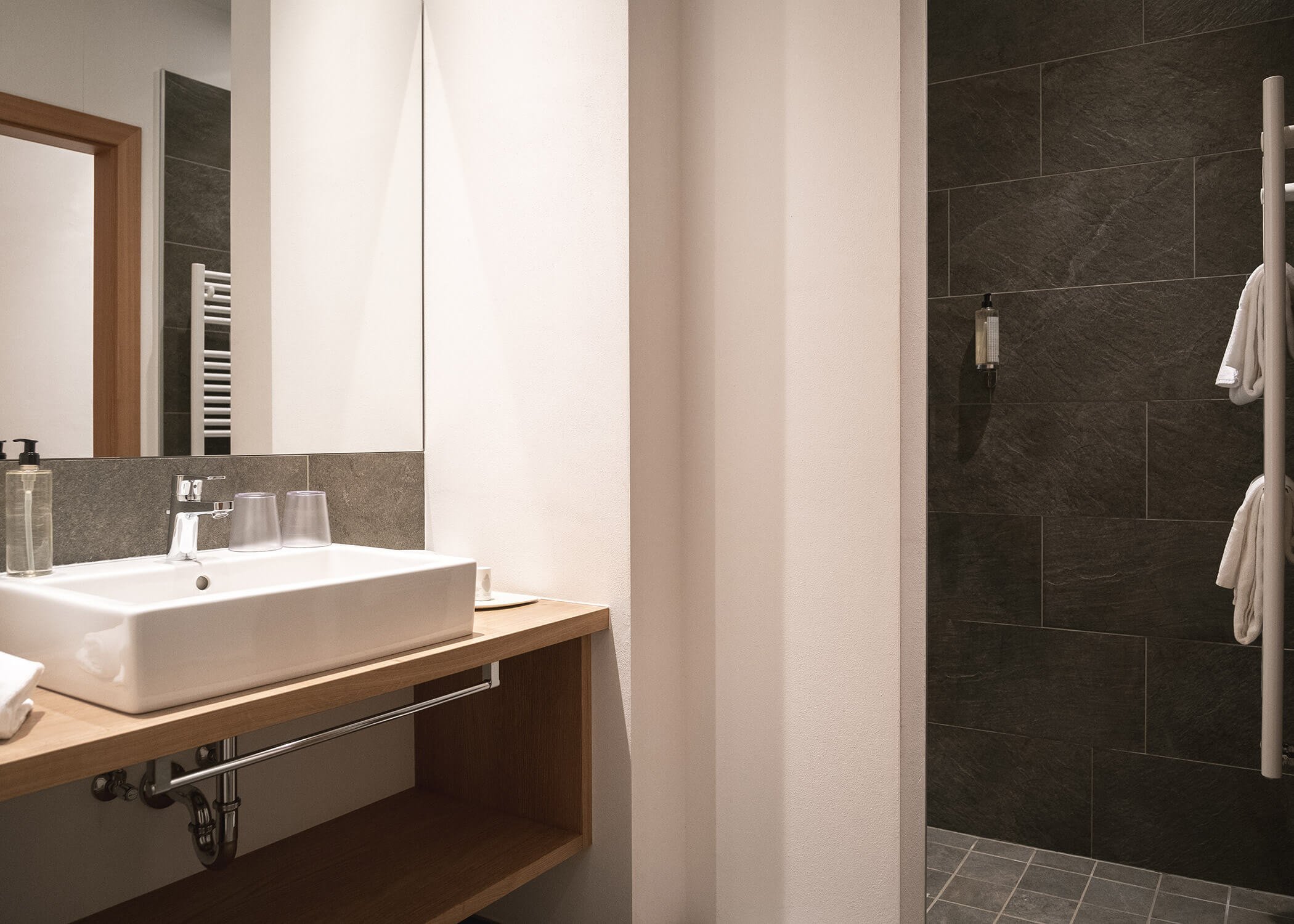 Modernes Badezimmer mit weißem Waschbecken, Holztisch, großem Spiegel und dunkel gefliestem Duschbereich. - Hotel La Casies Mountain Living