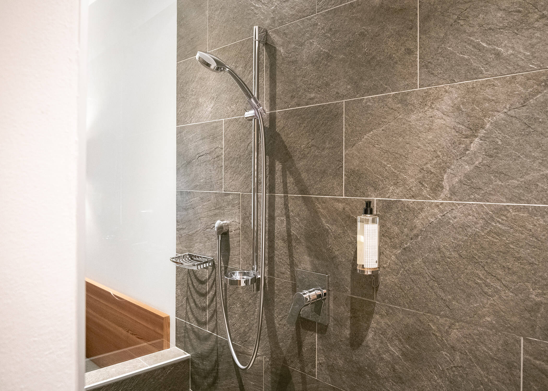 Moderne Dusche mit Handbrause, wandmontiertem Seifenspender und dunklen Steinfliesenwänden. - Hotel La Casies Mountain Living