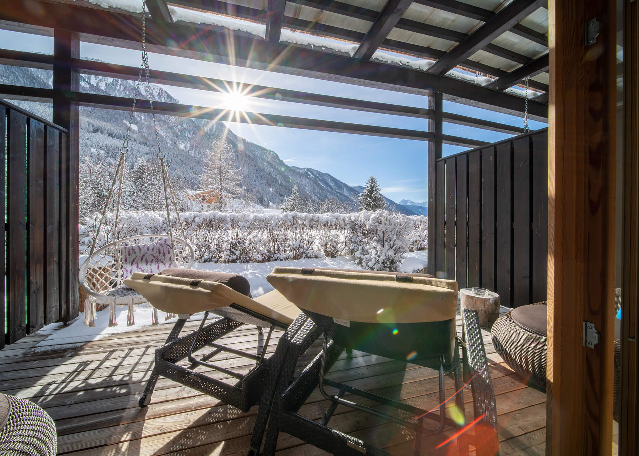Zwei Liegestühle auf einer Holzterrasse überblicken verschneite Berge und Bäume unter einem sonnigen, klaren Himmel. - Hotel La Casies Mountain Living