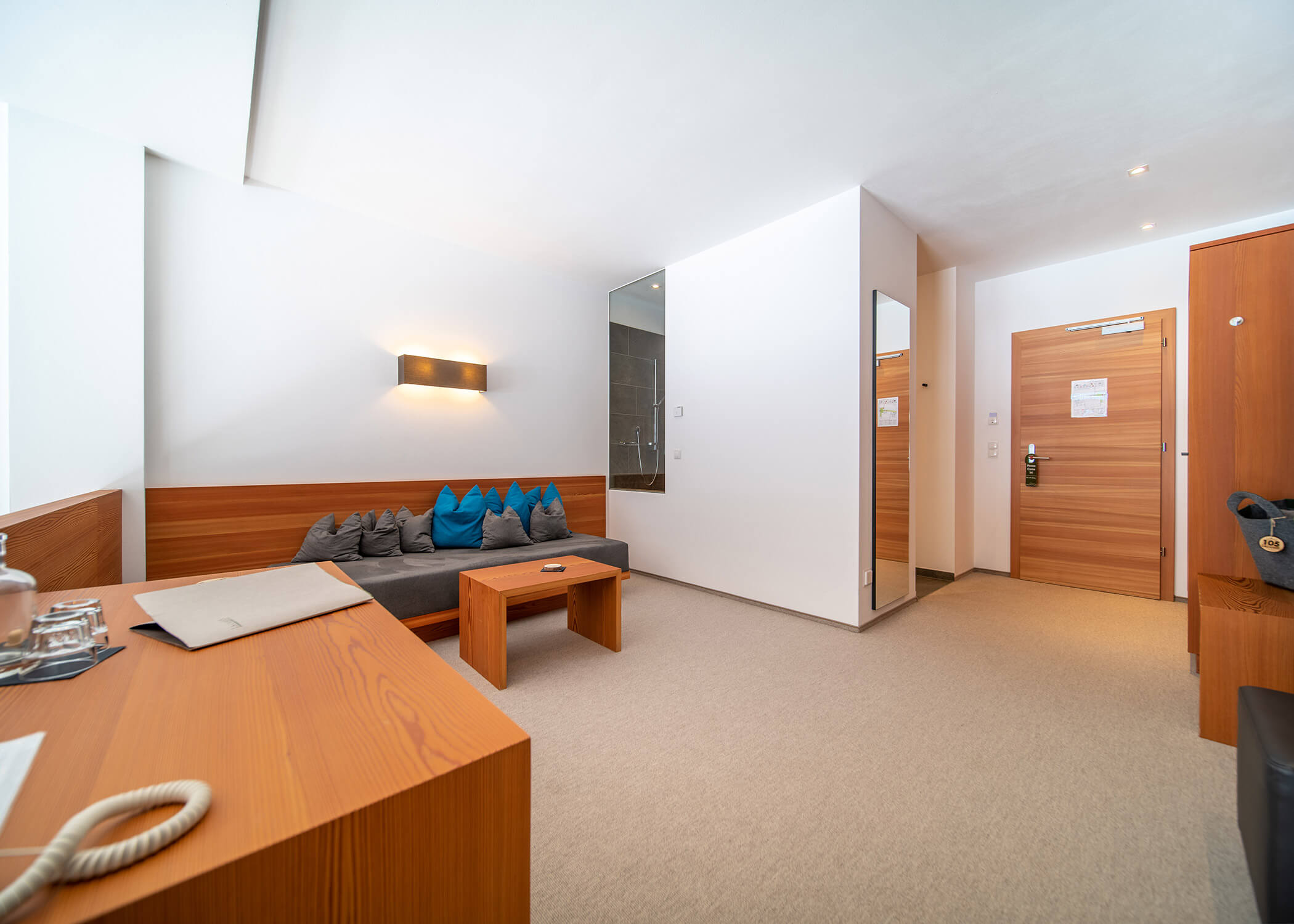 Modernes Hotelzimmer mit Holzmöbeln, einem grauen Sofa mit Kissen und einem kleinen Tisch neben der Eingangstür. - Hotel La Casies Mountain Living