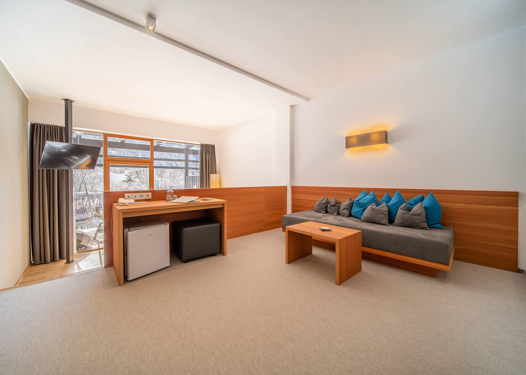 Minimalistisches Hotelzimmer mit einem Schreibtisch, einem kleinen Kühlschrank, einem langen Sofa mit blauen Kissen und einem Fenster mit Vorhängen. - Hotel La Casies Mountain Living