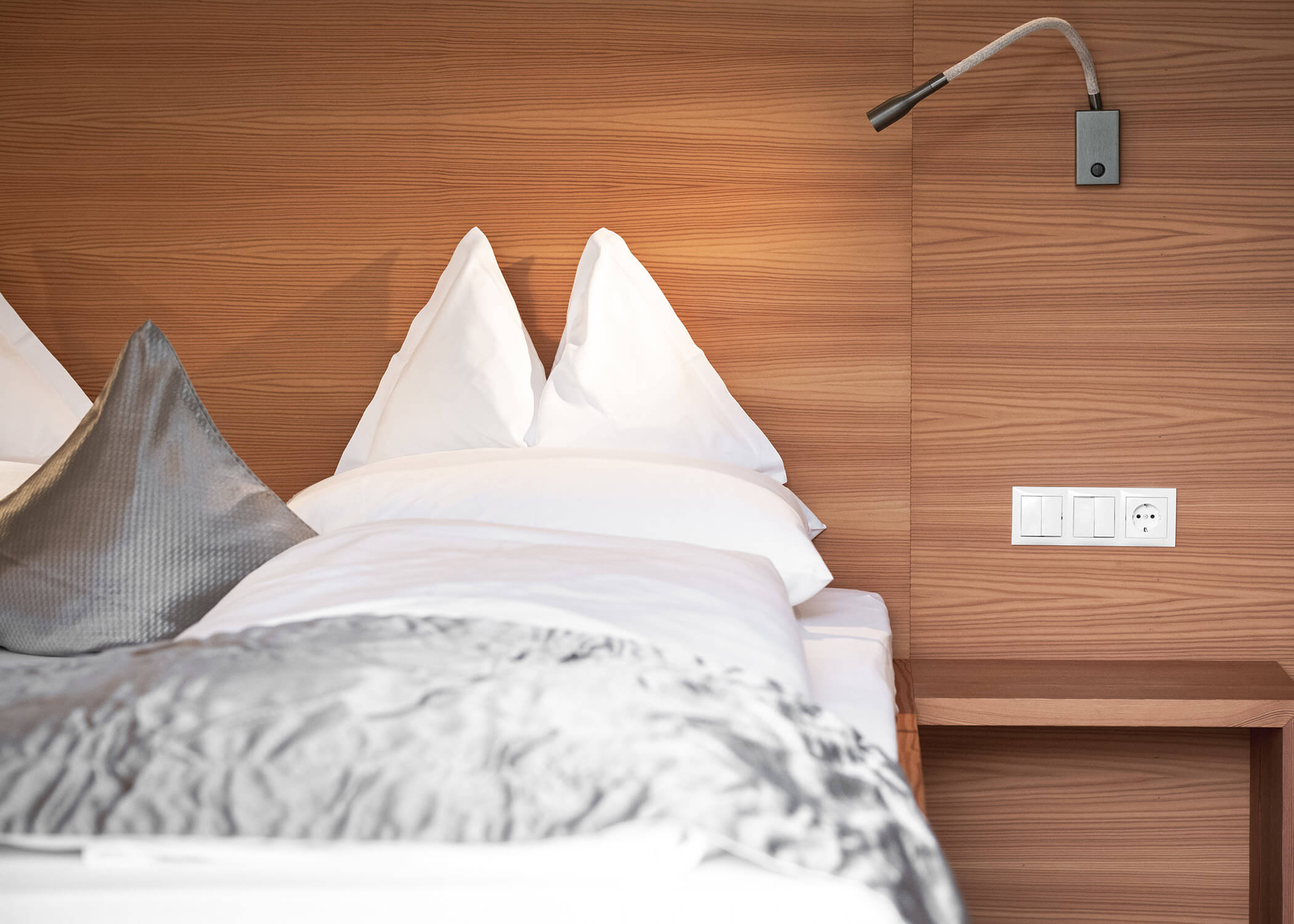 Modernes Hotelbett mit weißer Bettwäsche, grauer Decke, Leselicht und Holzwand mit Steckdosen. - Hotel La Casies Mountain Living