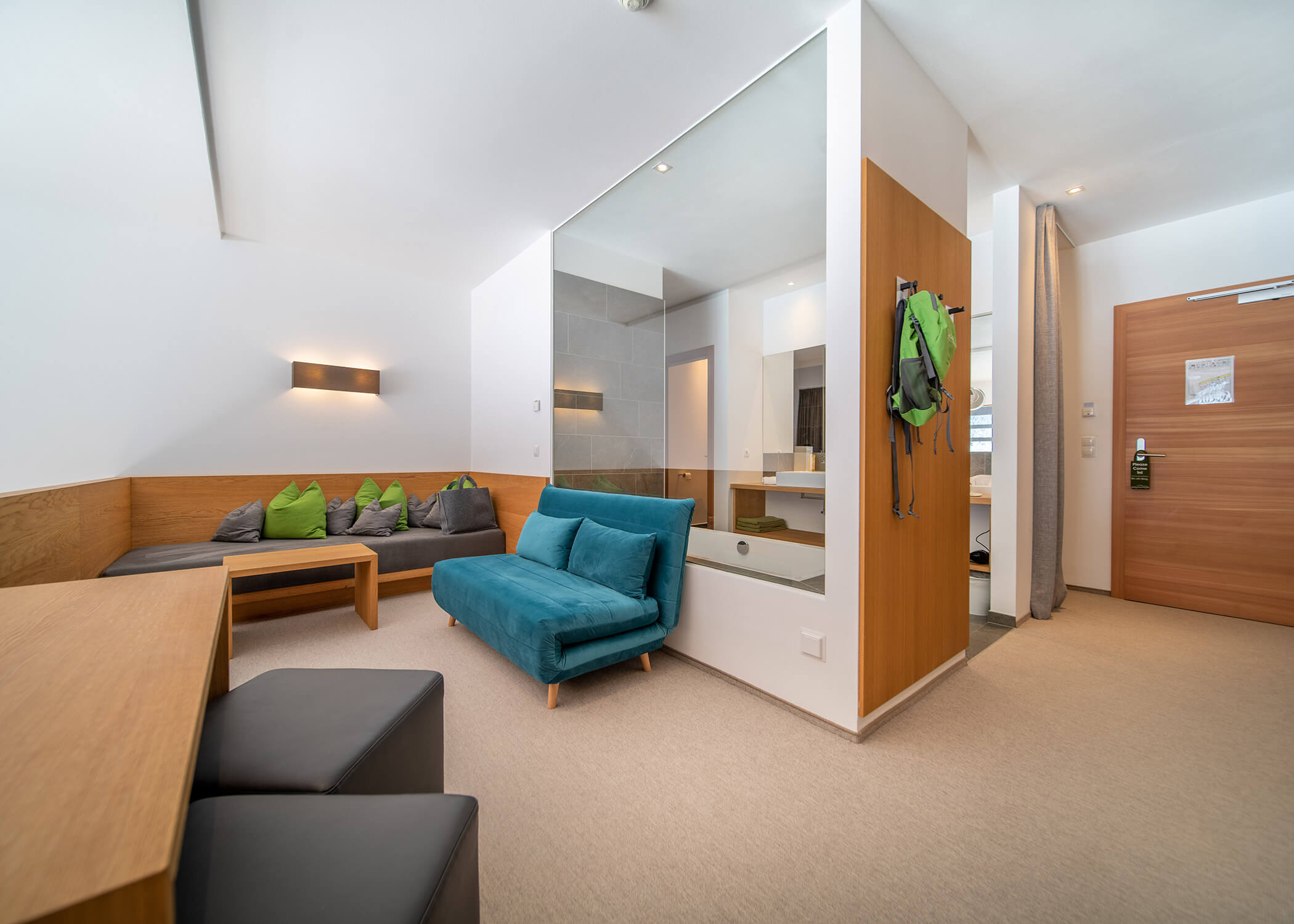 Modernes Hotelzimmer mit blauem Sofa, Sitzecke und grünem Rucksack, der an einem Glastrenner neben dem Badezimmer hängt. - Hotel La Casies Mountain Living