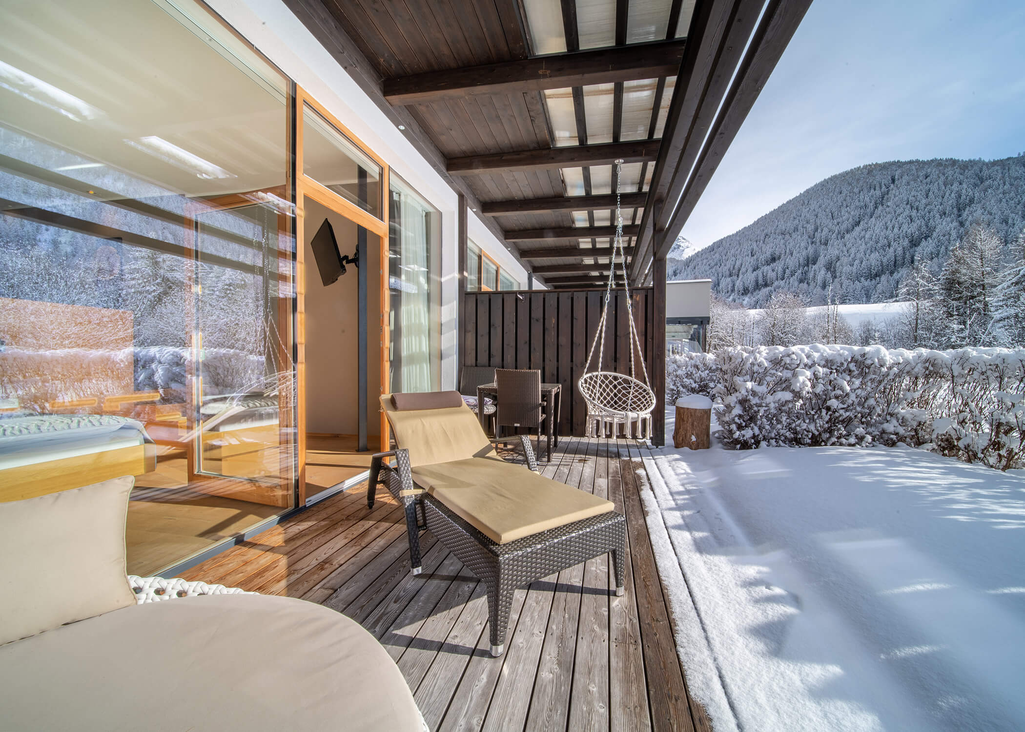 Sonnige Holzterrasse mit Liegestühlen, einer Schaukel und schneebedeckter Landschaft mit Bergen im Hintergrund. - Hotel La Casies Mountain Living
