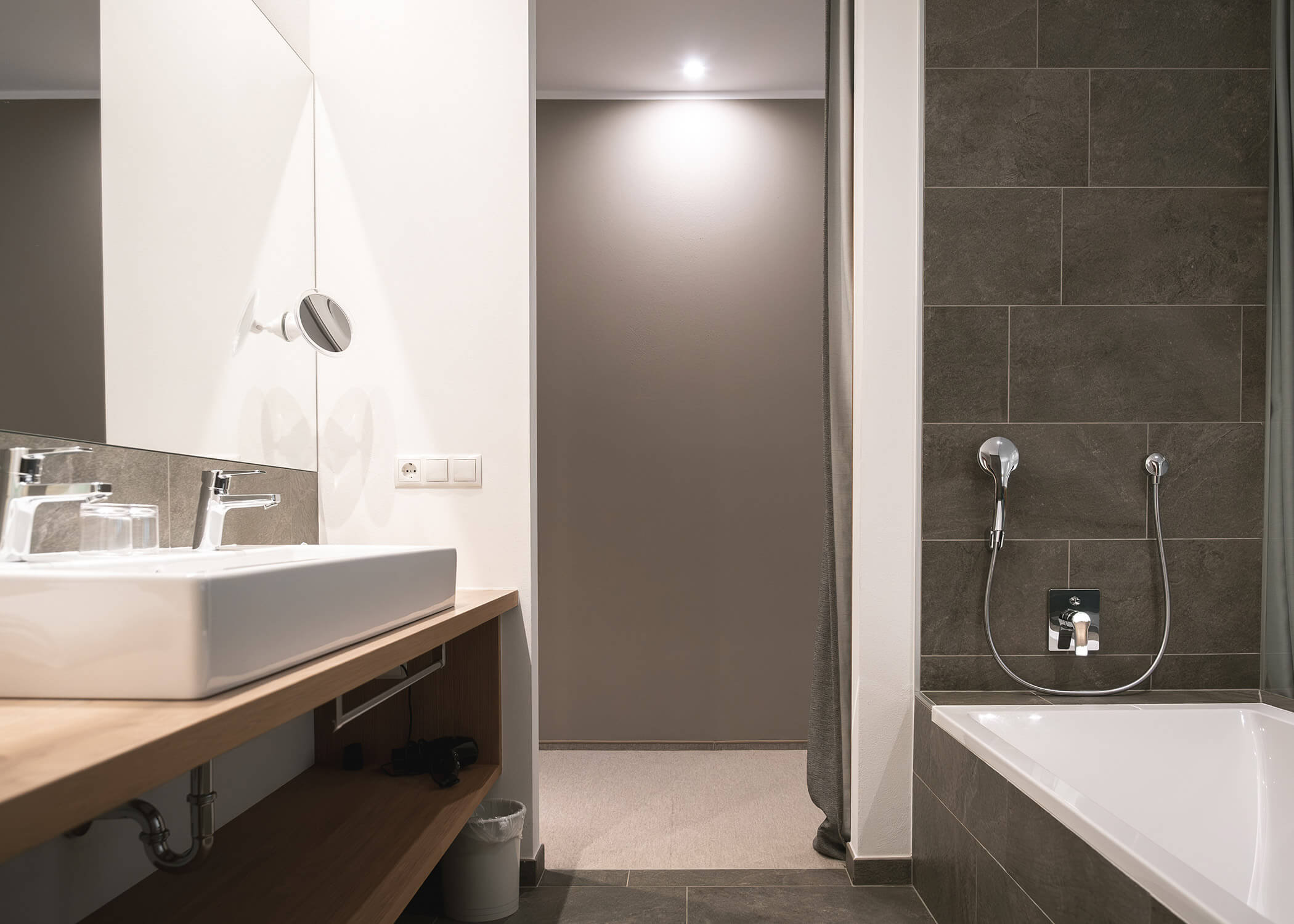 Modernes Badezimmer mit weißem Waschbecken, Spiegel und Badewanne, mit grauen Fliesen und neutralen Wänden. - Hotel La Casies Mountain Living