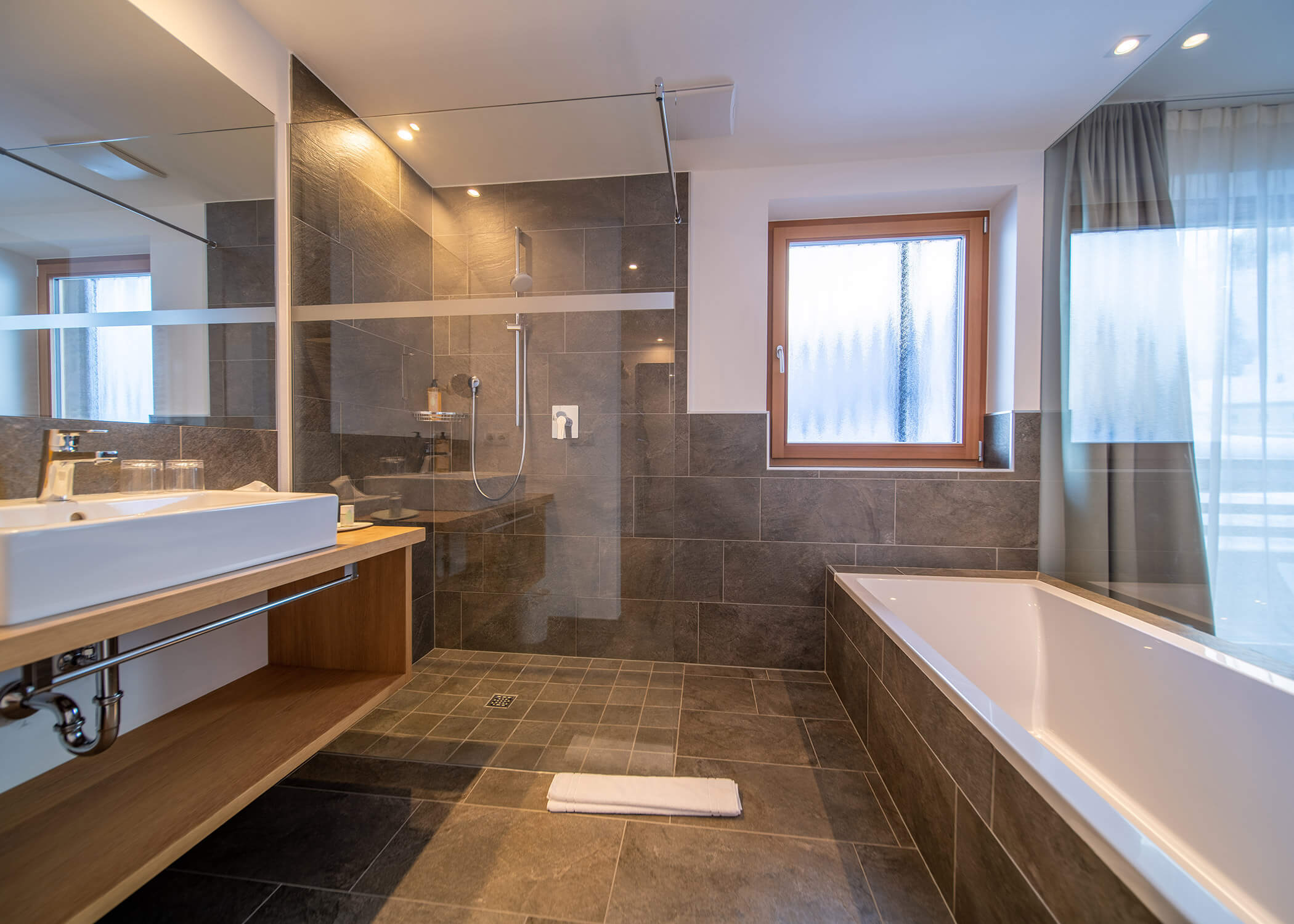 Modernes Badezimmer mit grauen Fliesen, begehbarer Glasdusche, Badewanne, Waschbecken und Milchglasfenstern. - Hotel La Casies Mountain Living