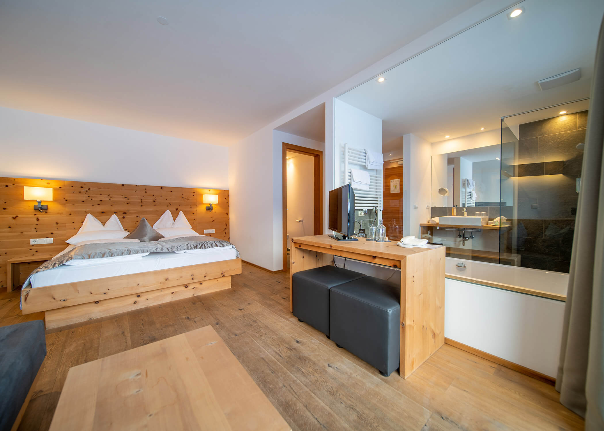 Modernes Hotelzimmer mit Holzmöbeln, Doppelbett, Schreibtisch, TV und offenem Bad mit Glasdusche. - Hotel La Casies Mountain Living