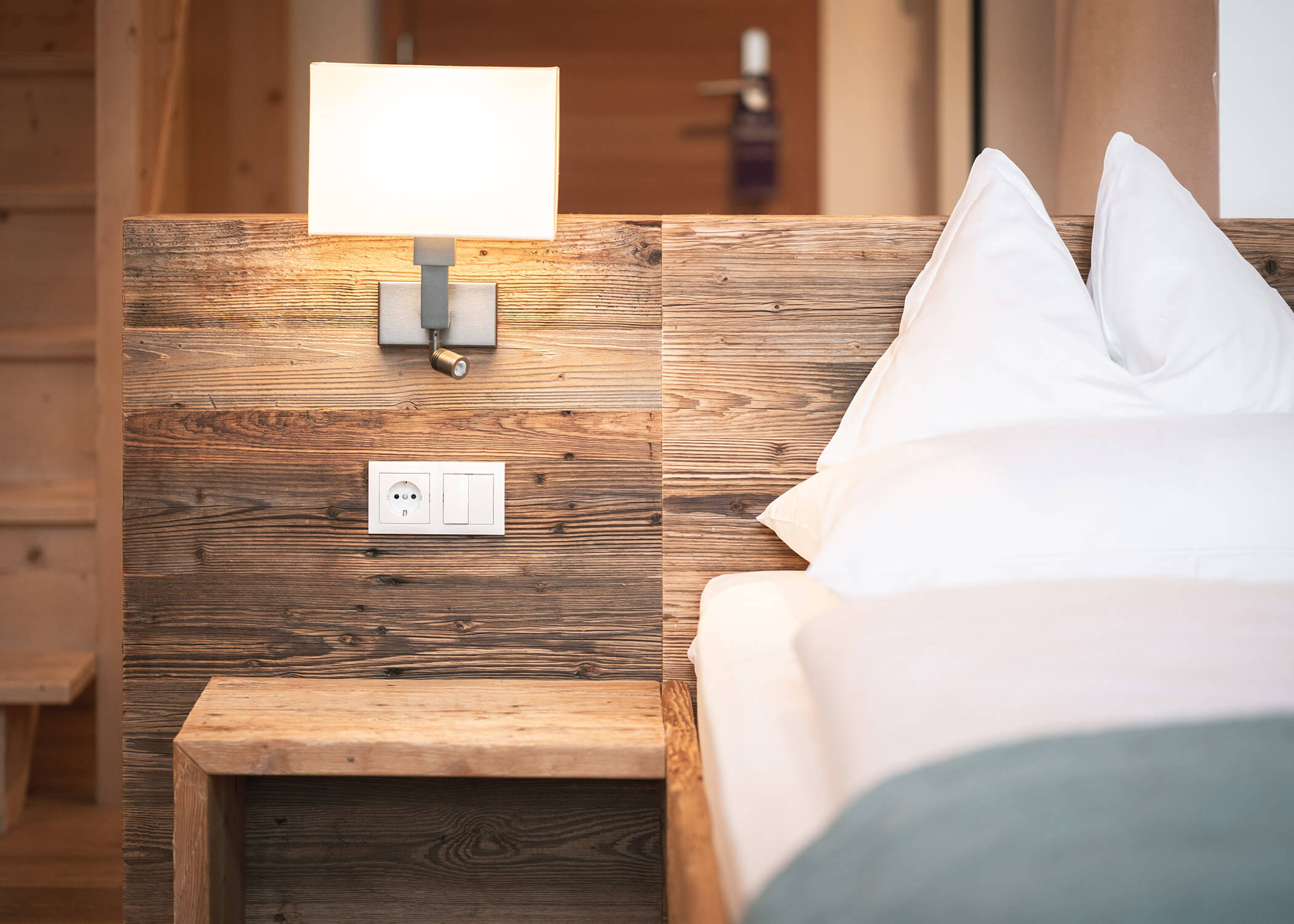 Bett mit weißer Bettwäsche neben einem hölzernen Nachttisch, Wandlampe und Steckdose in einem gemütlichen Zimmer. - Hotel La Casies Mountain Living