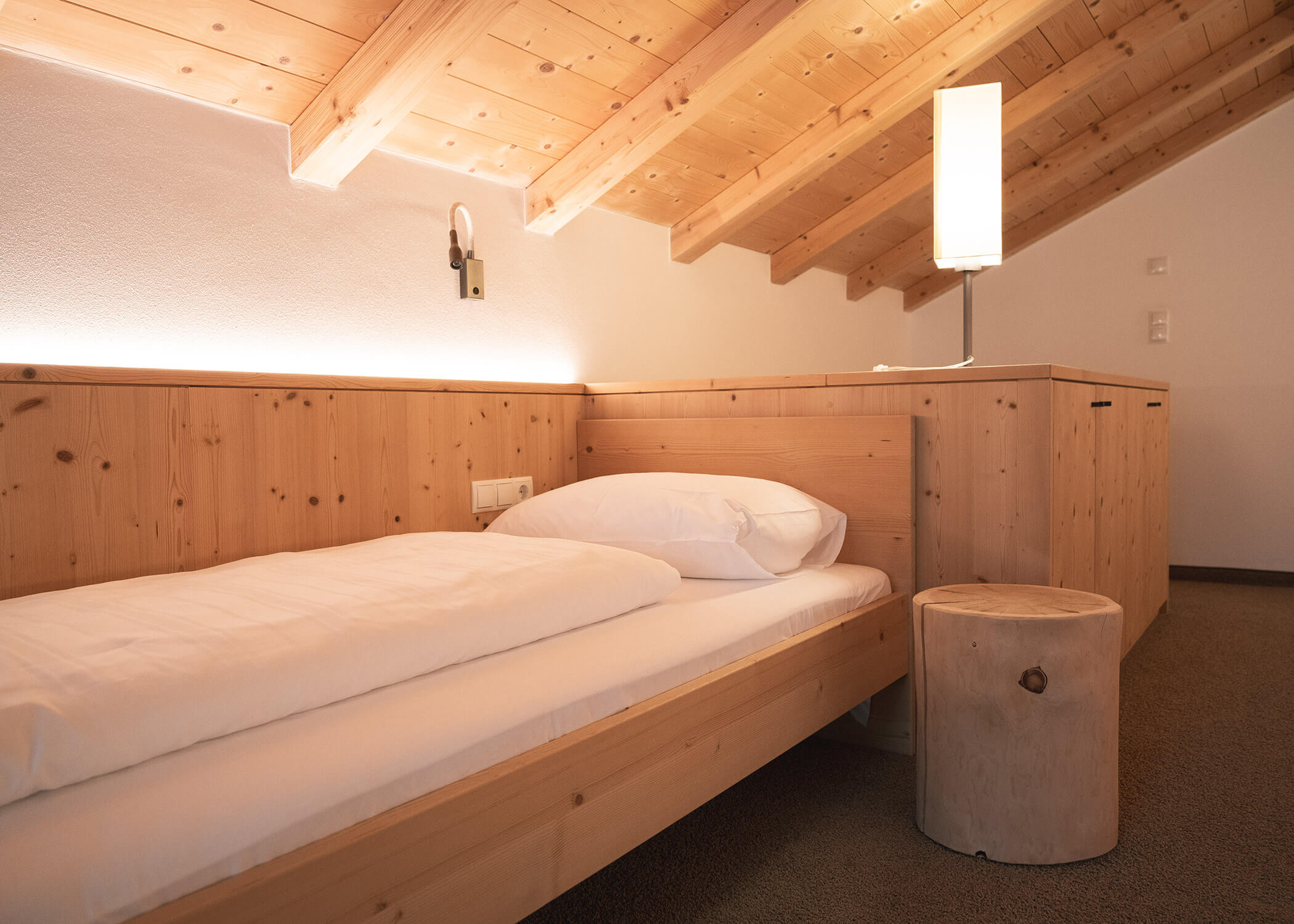 Ein gemütliches Dachzimmer aus Holz mit einem Einzelbett, einer Lampe und einem rustikalen Hocker unter einer schrägen Decke. - Hotel La Casies Mountain Living