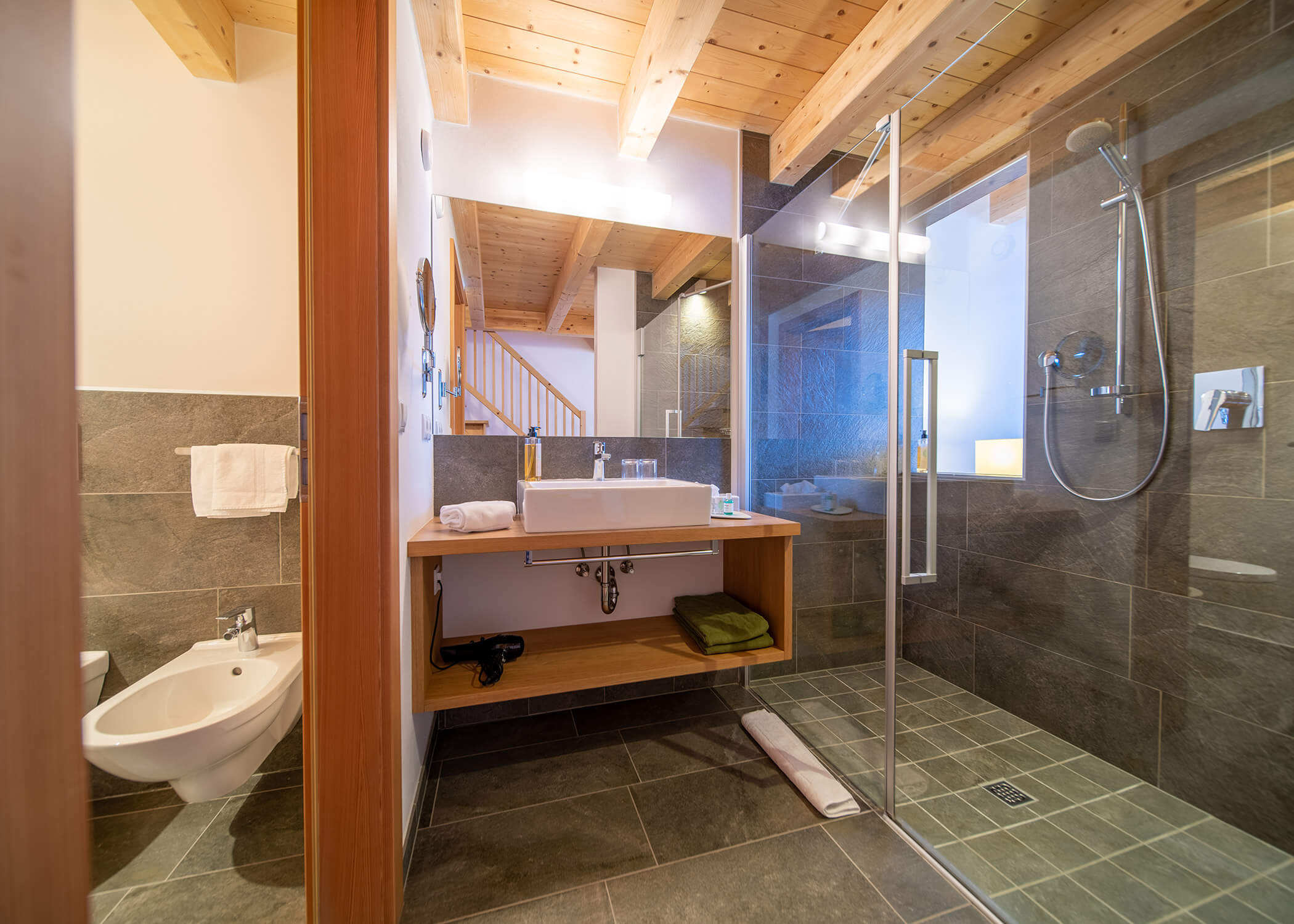 Modernes Badezimmer mit Glasdusche, Bidet, Holzdecke und großem Spiegel über einem Waschbecken mit Handtüchern darunter. - Hotel La Casies Mountain Living