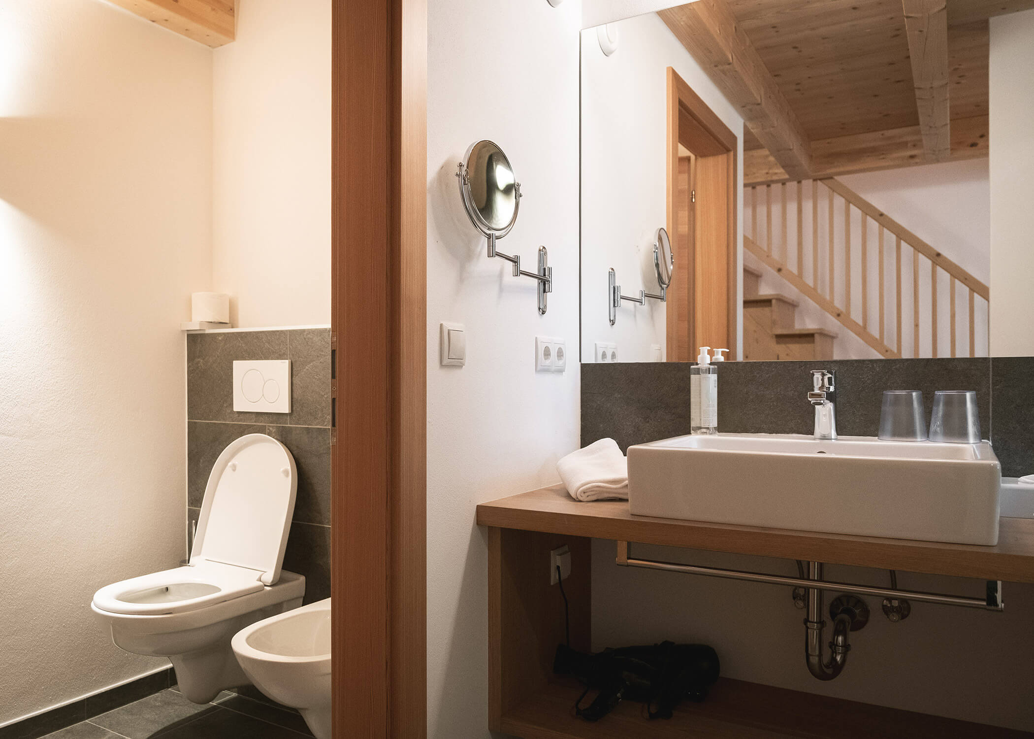 Modernes Badezimmer mit Toilette, Bidet, Waschbecken, Spiegel und hölzernen Akzenten, mit einer sichtbaren Treppe im Hintergrund. - Hotel La Casies Mountain Living